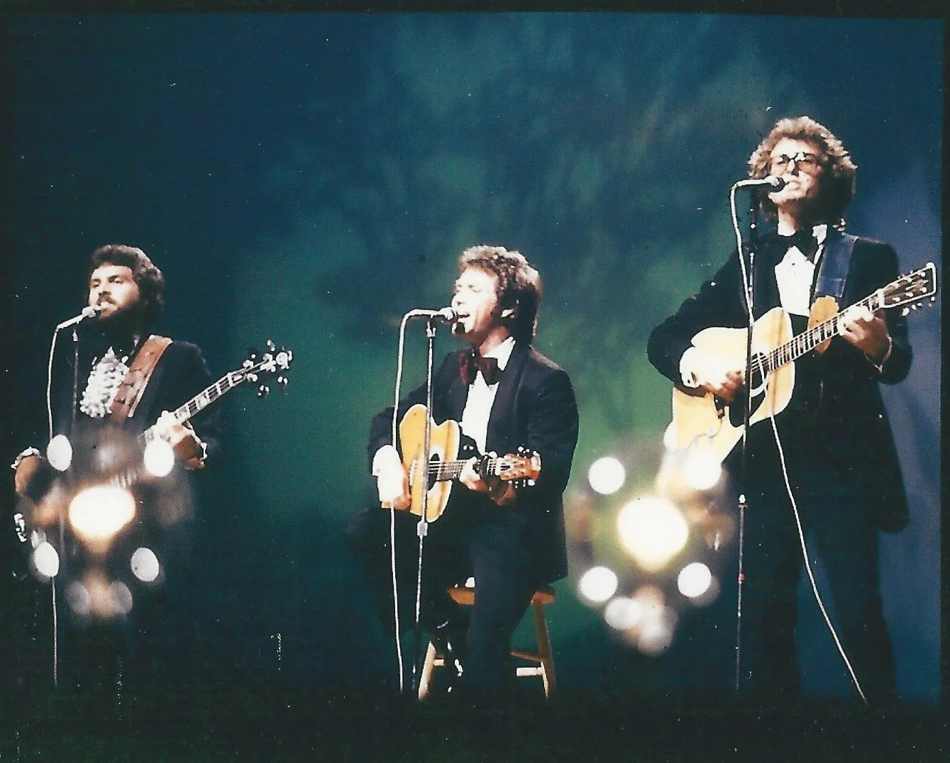 Three musicians performing on stage, each playing an acoustic guitar and singing into a microphone. The background is dark with out-of-focus stage lights.
