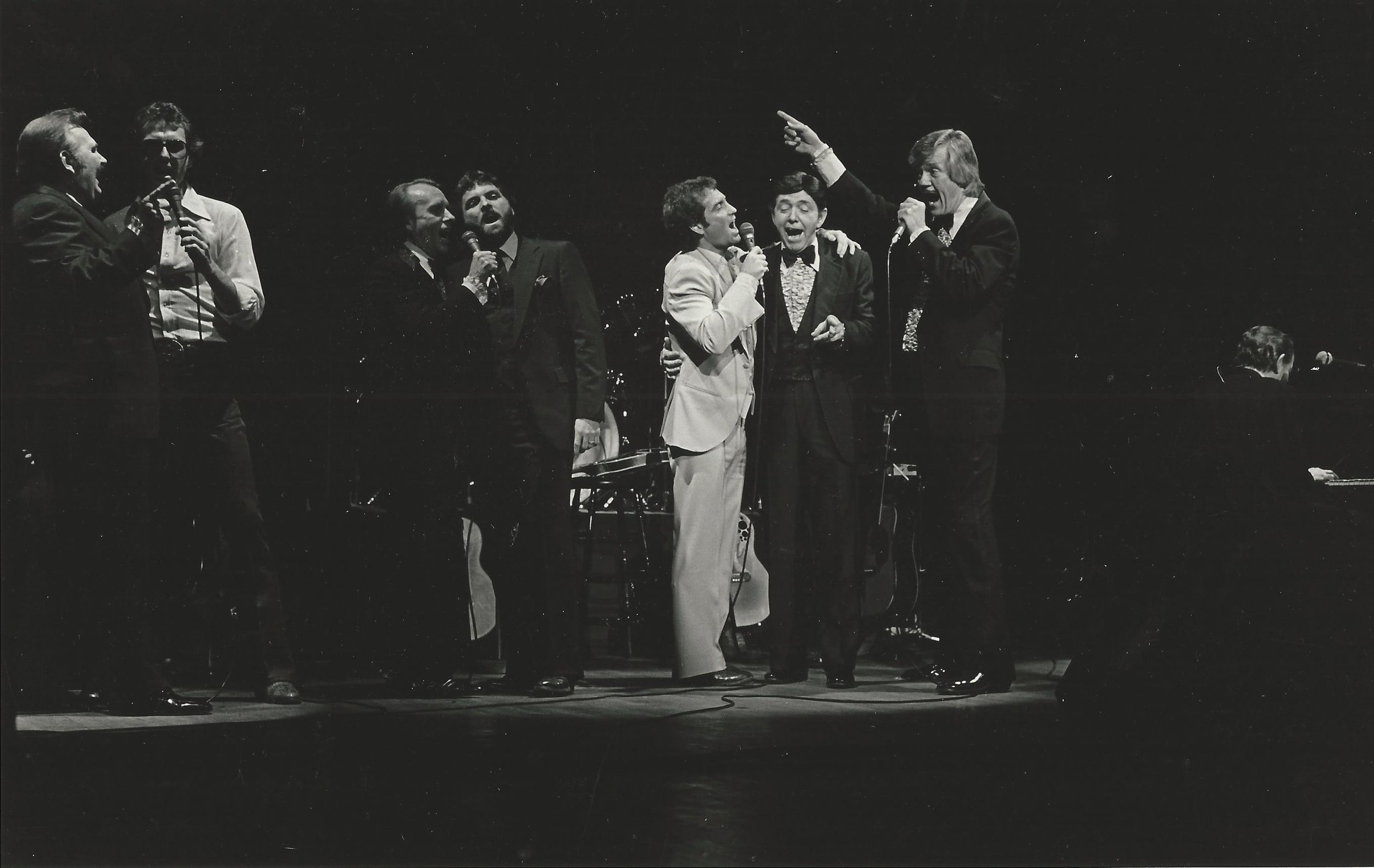Black and white photo of seven performers singing and engaging with each other on stage. They are holding microphones, dressed in formal and stylish clothing, with one person enthusiastically pointing upward.