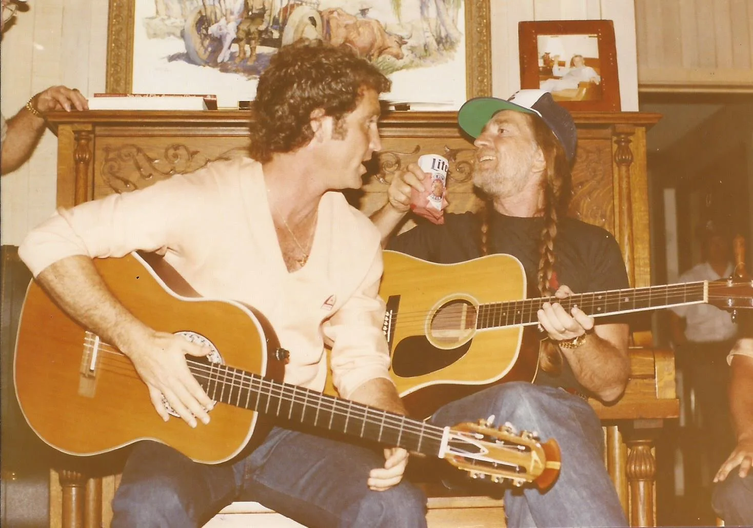 Two men, Larry Gatlin and Willie Nelson, playing guitars and singing together in a cozy room, one wearing a light-colored shirt with rolled-up sleeves and the other wearing a black shirt, a hat, and braids, with a person reaching with a beverage.