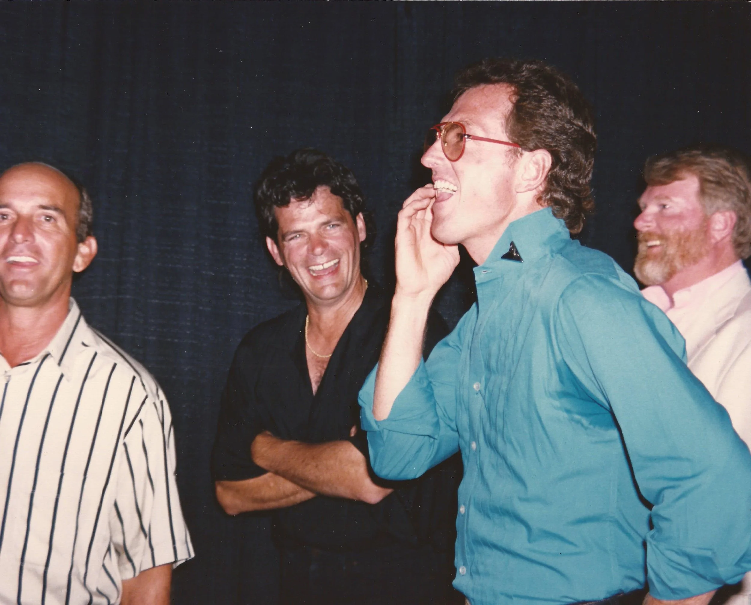Group of people laughing and smiling, one person with sunglasses and a bright blue shirt, at a social gathering.