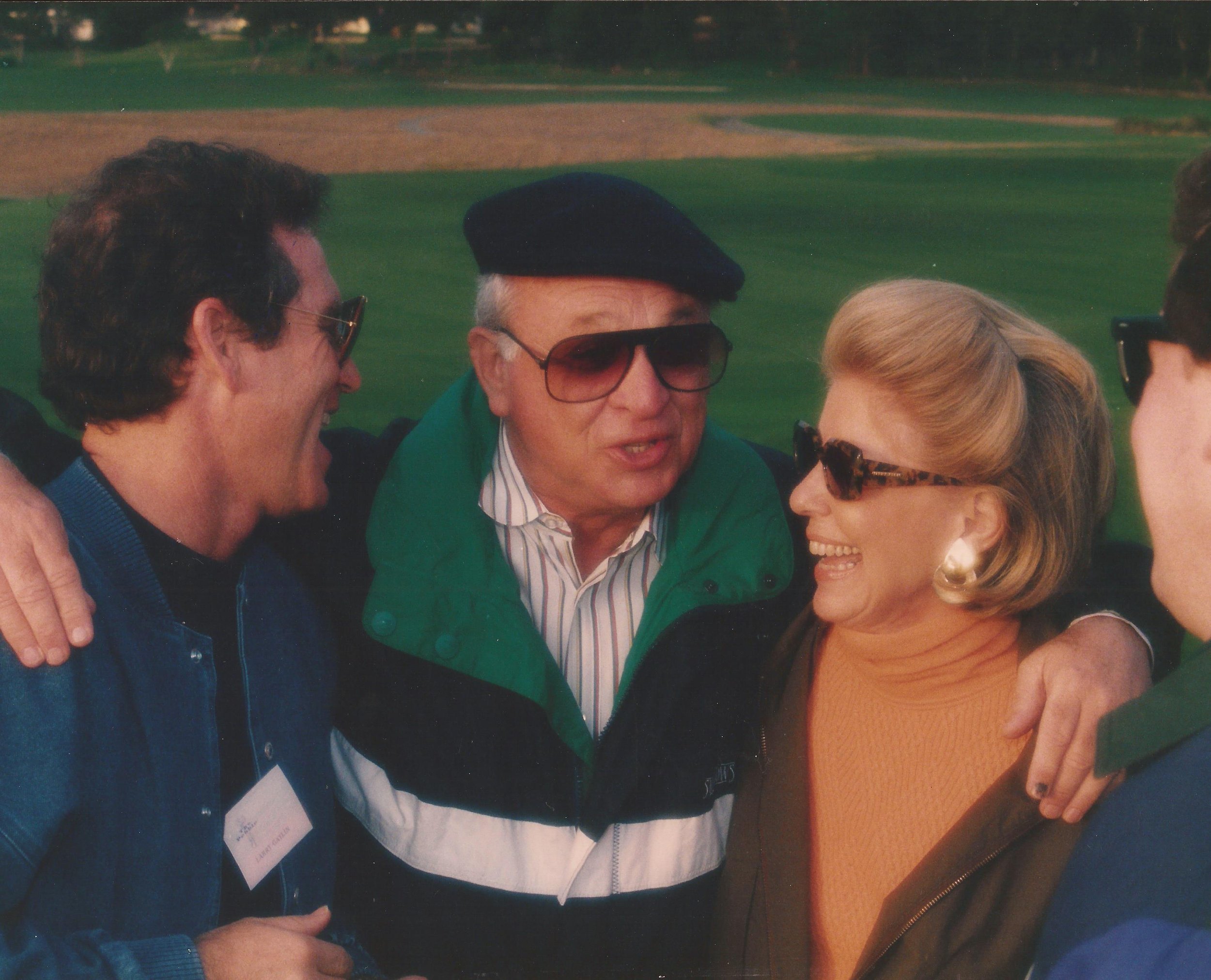People in a social gathering, smiling, talking, and wearing casual clothing outdoors, with a green, field-like background.