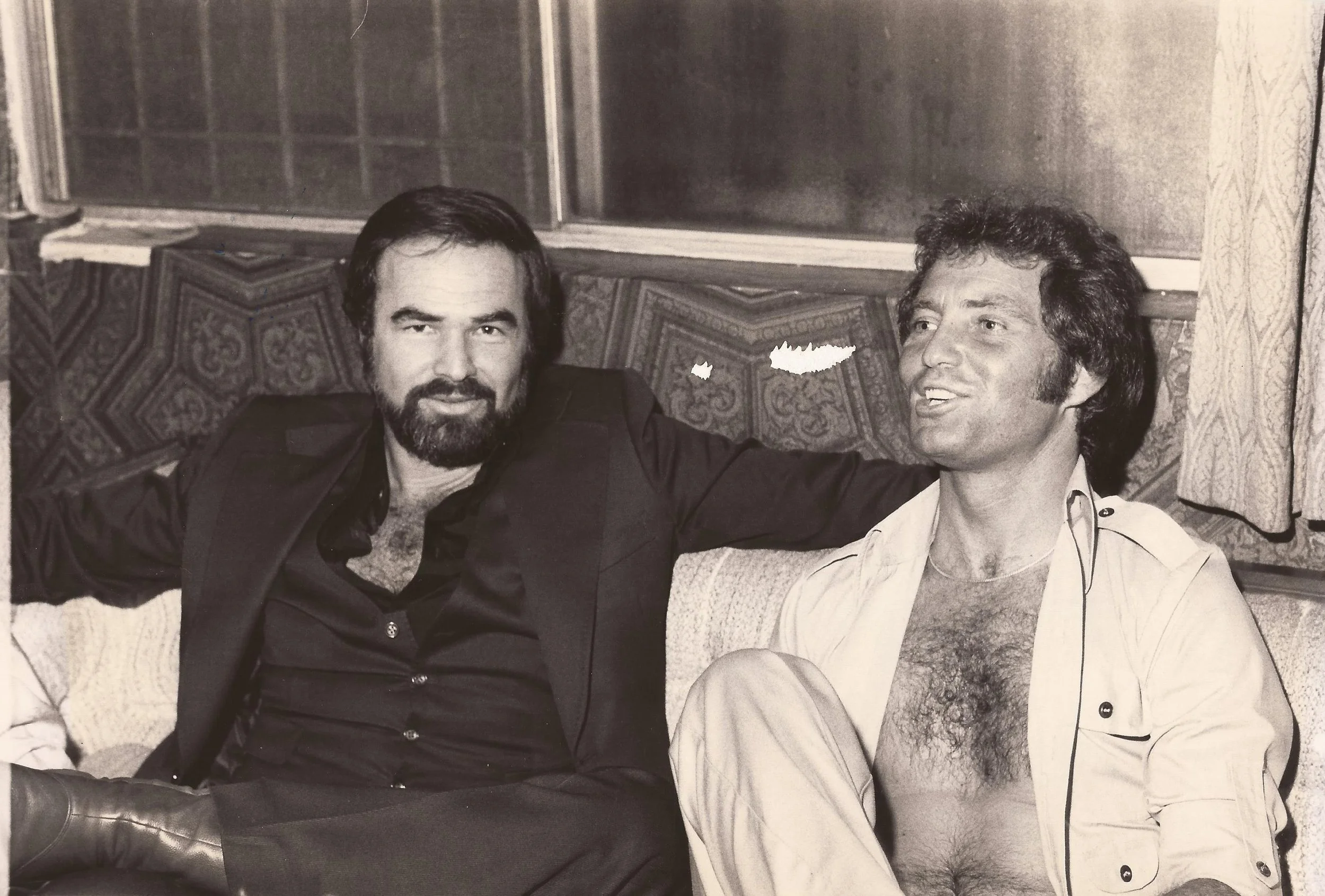 Two men sitting on a patterned bench in front of a window, smiling and relaxed, in a black-and-white photo.