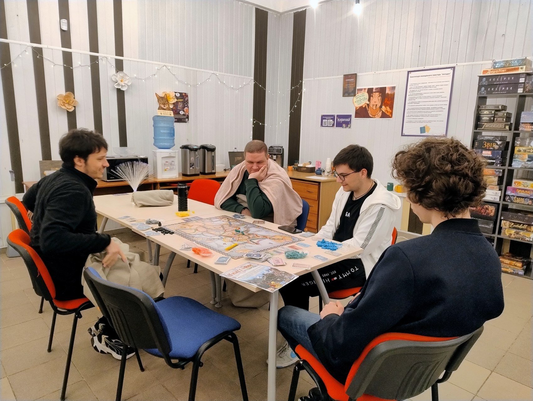 Four people sitting around a table playing a board game, with game pieces and cards on the table.