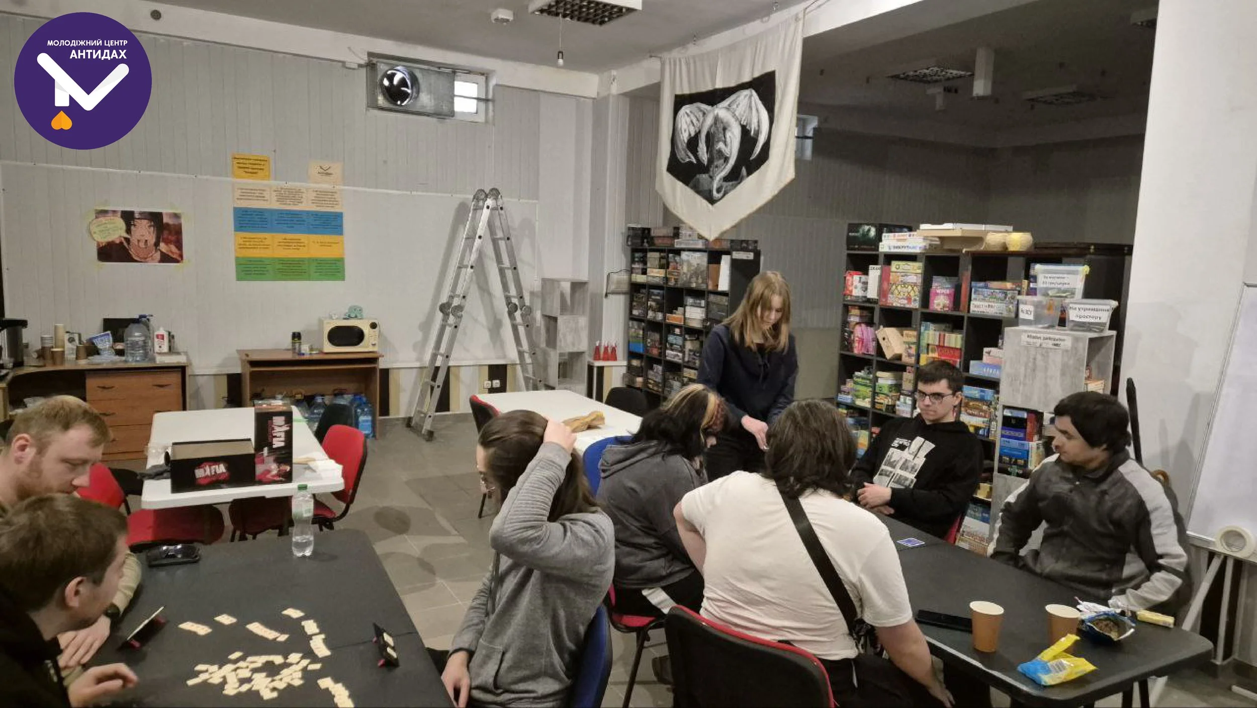People gathered around tables playing board games in a room filled with bookshelves, posters, and a ladder hanging on the wall.
