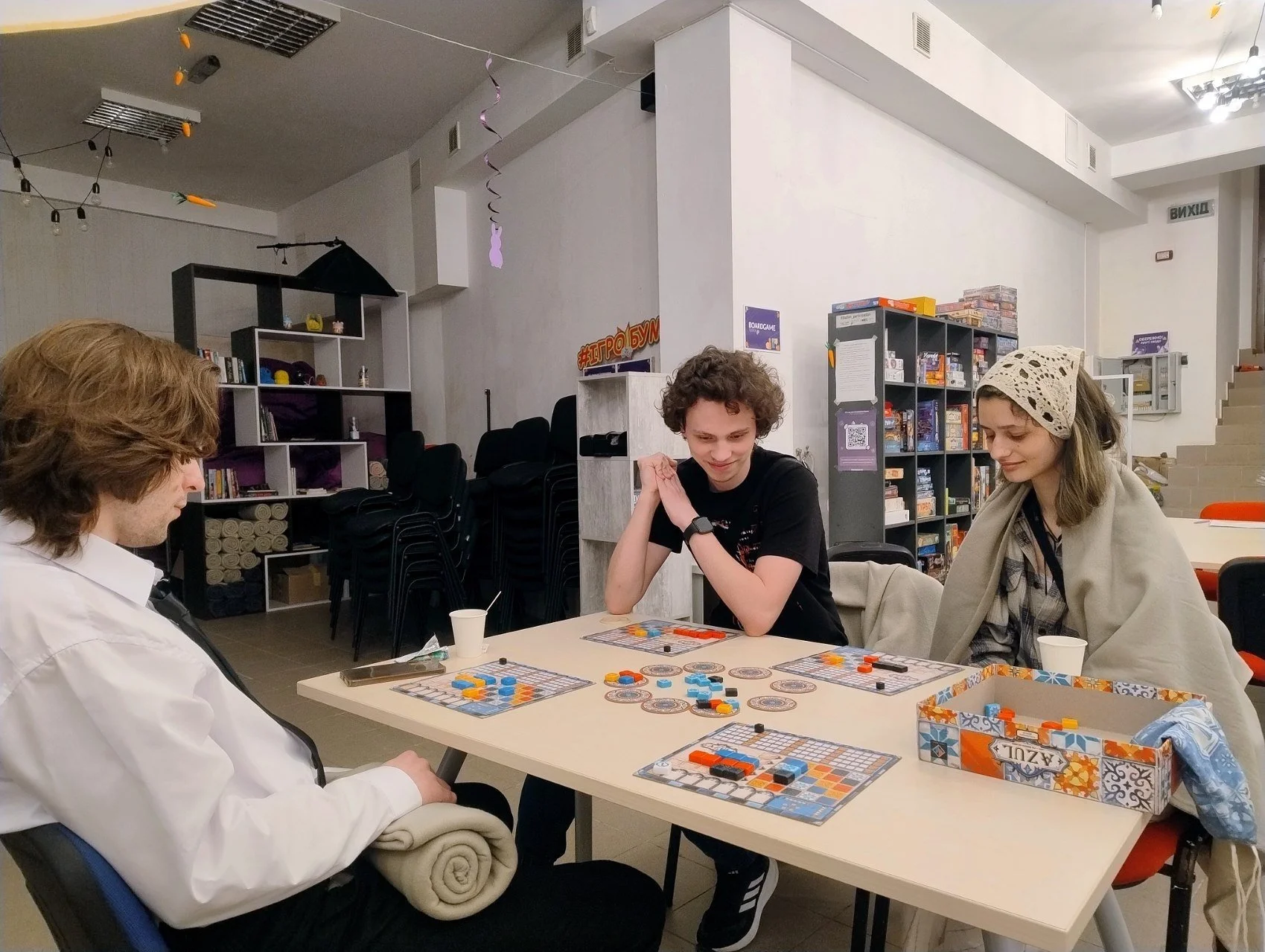 Three young adults playing a board game at a table in a well-lit room with storage shelves and board games in the background.