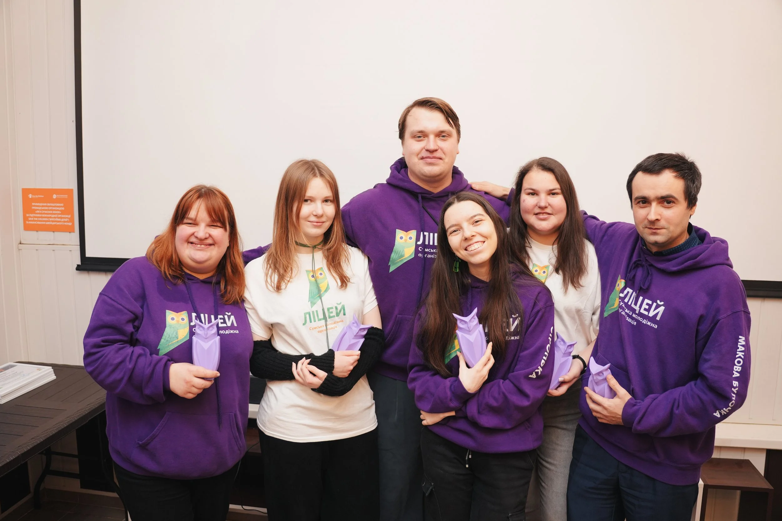 Group of five young people standing together indoors, smiling and holding purple paper owl-shaped objects, some wearing purple hoodies and one in white shirt, in front of a whiteboard or screen.
