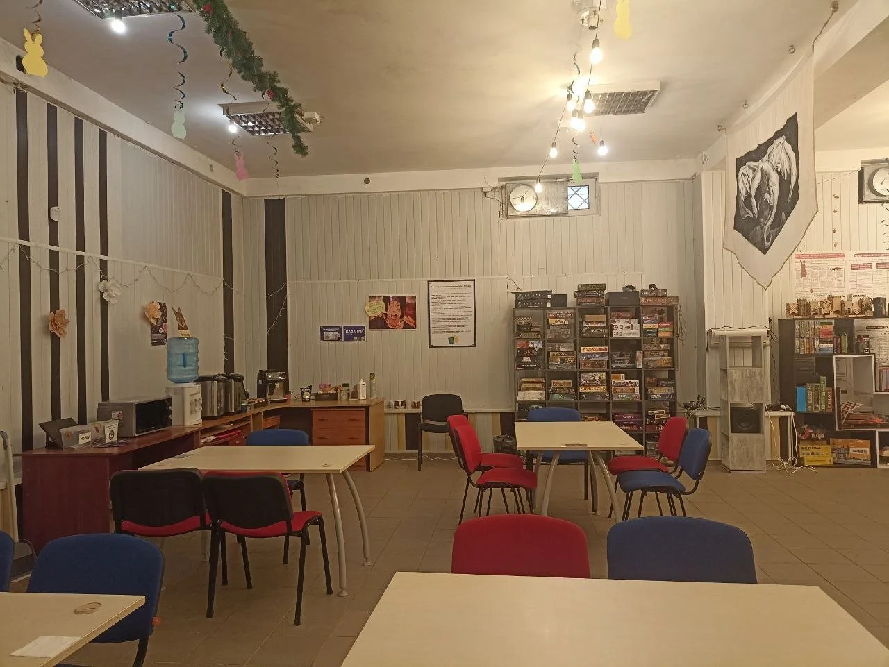 An empty room with tables and chairs, decorated with hanging paper cutouts and garlands, featuring a water dispenser, shelves with board games, and posters on the walls.