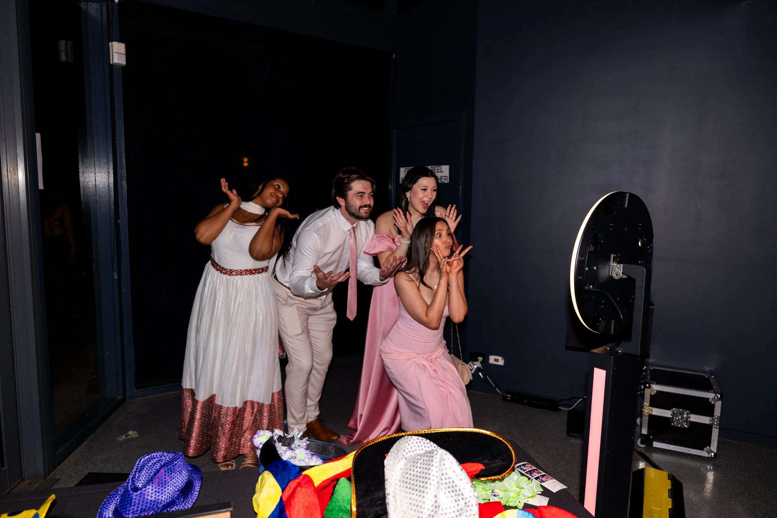 Group of five people in formal attire taking a fun photo with a photo booth, making playful expressions, with props and photo booth equipment in the foreground.