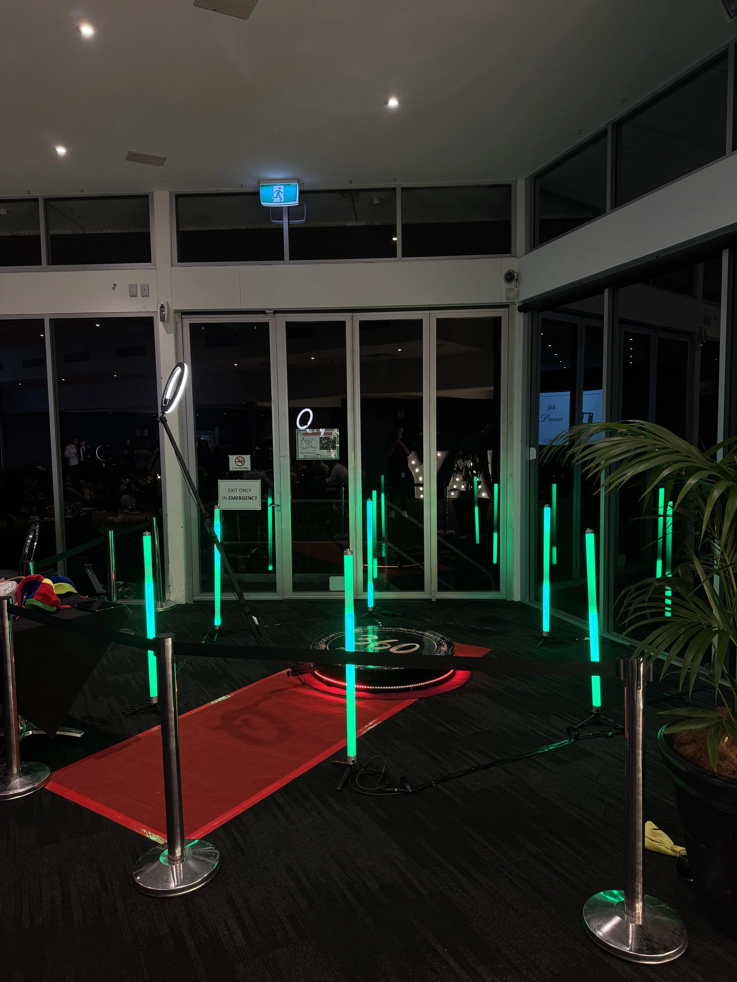 A virtual golf simulator setup with a red carpet, green LED light posts, and a hitting surface in an indoor space with glass doors and windows.