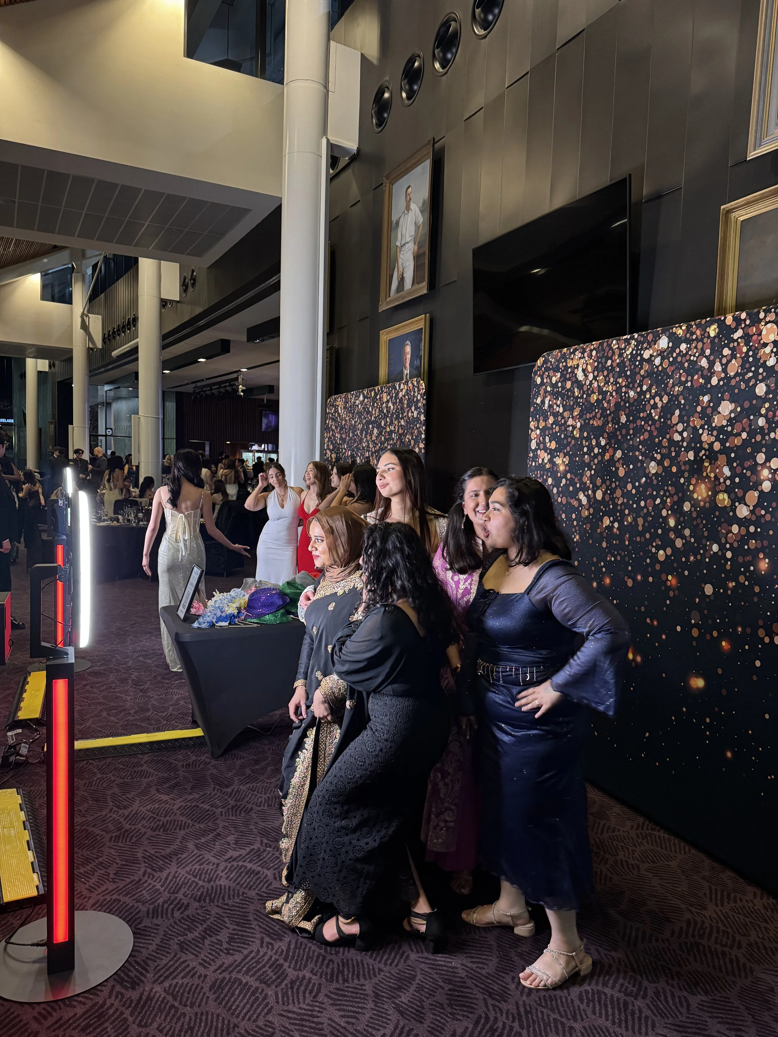 Group of women posing for a photo at an indoor event, with a dark wall decorated with paintings and a patterned backdrop behind them.