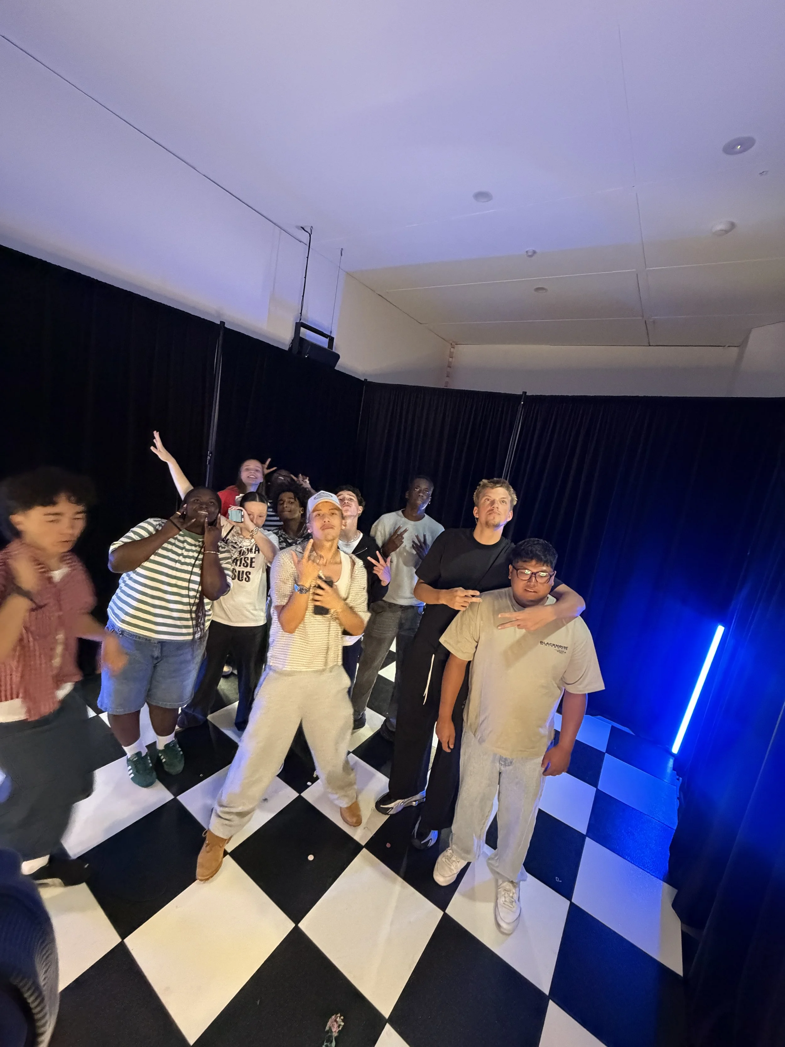 Group of young people posing and having fun on a checkered dance floor with black curtains and a vertical blue neon light in the background.