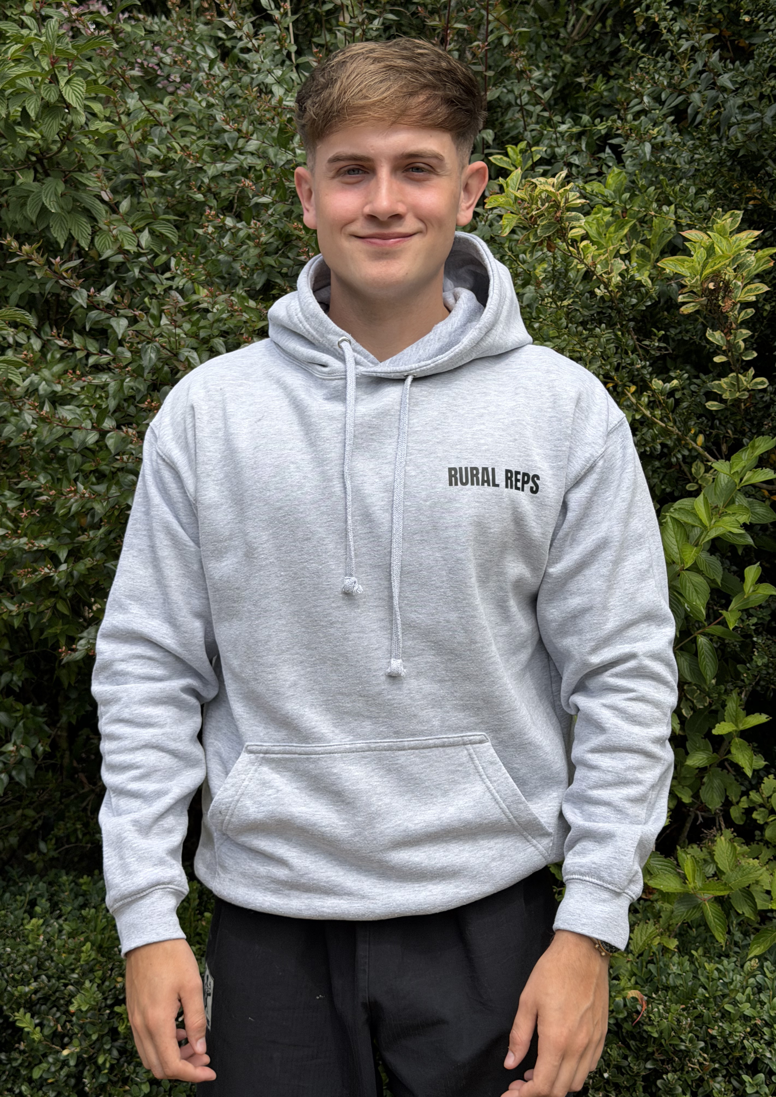 A young man with light skin, short brown hair, and blue eyes smiling slightly in front of green foliage, wearing a light gray hoodie with black text that reads 'RURAL REPS'.