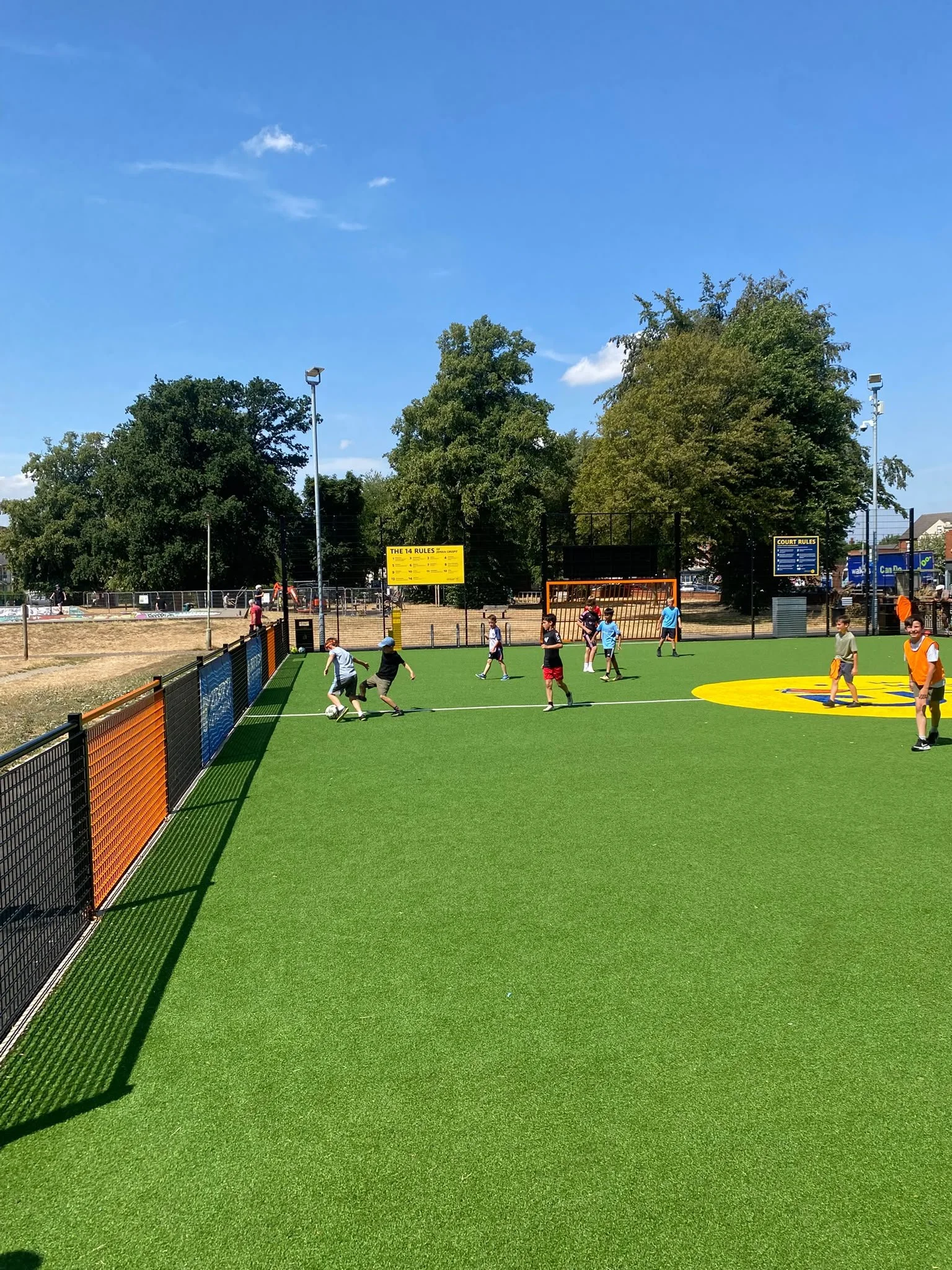 Children playing soccer on a small artificial turf field enclosed by a black fence, with trees, light poles, and a bright blue sky in the background.