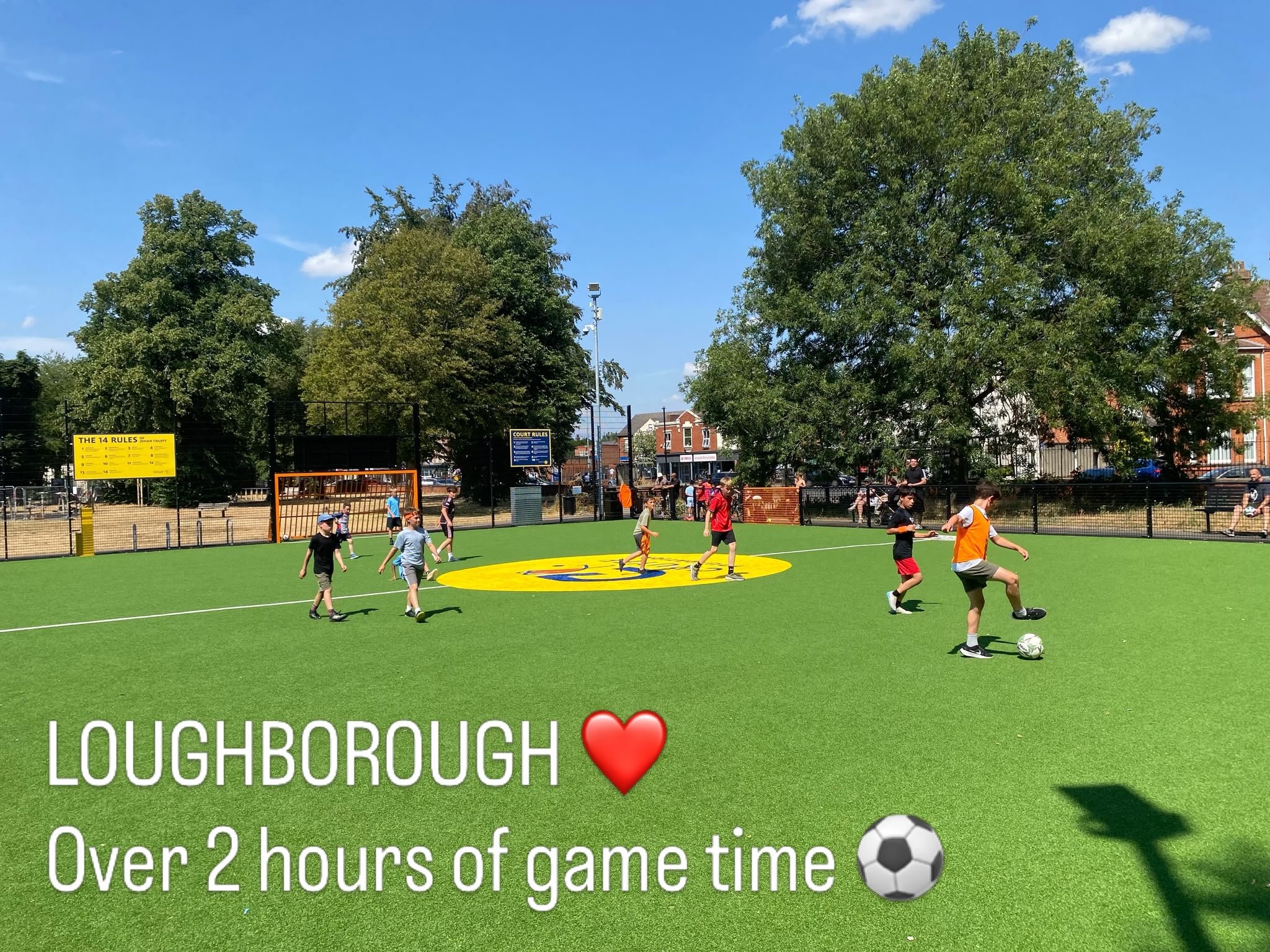 Children playing soccer on a green field in a neighborhood park with tall trees, buildings, and a blue sky in the background, and a sign with rules.