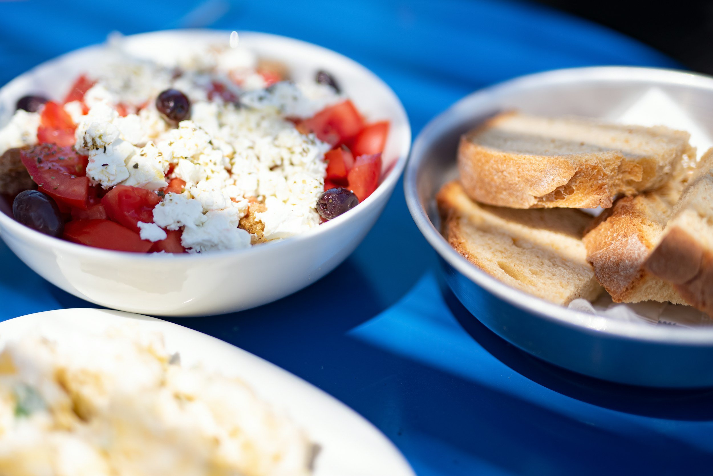 Bowl of Greek salad with tomatoes, black olives, feta cheese, and cucumbers, next to a bowl of sliced bread with butter.