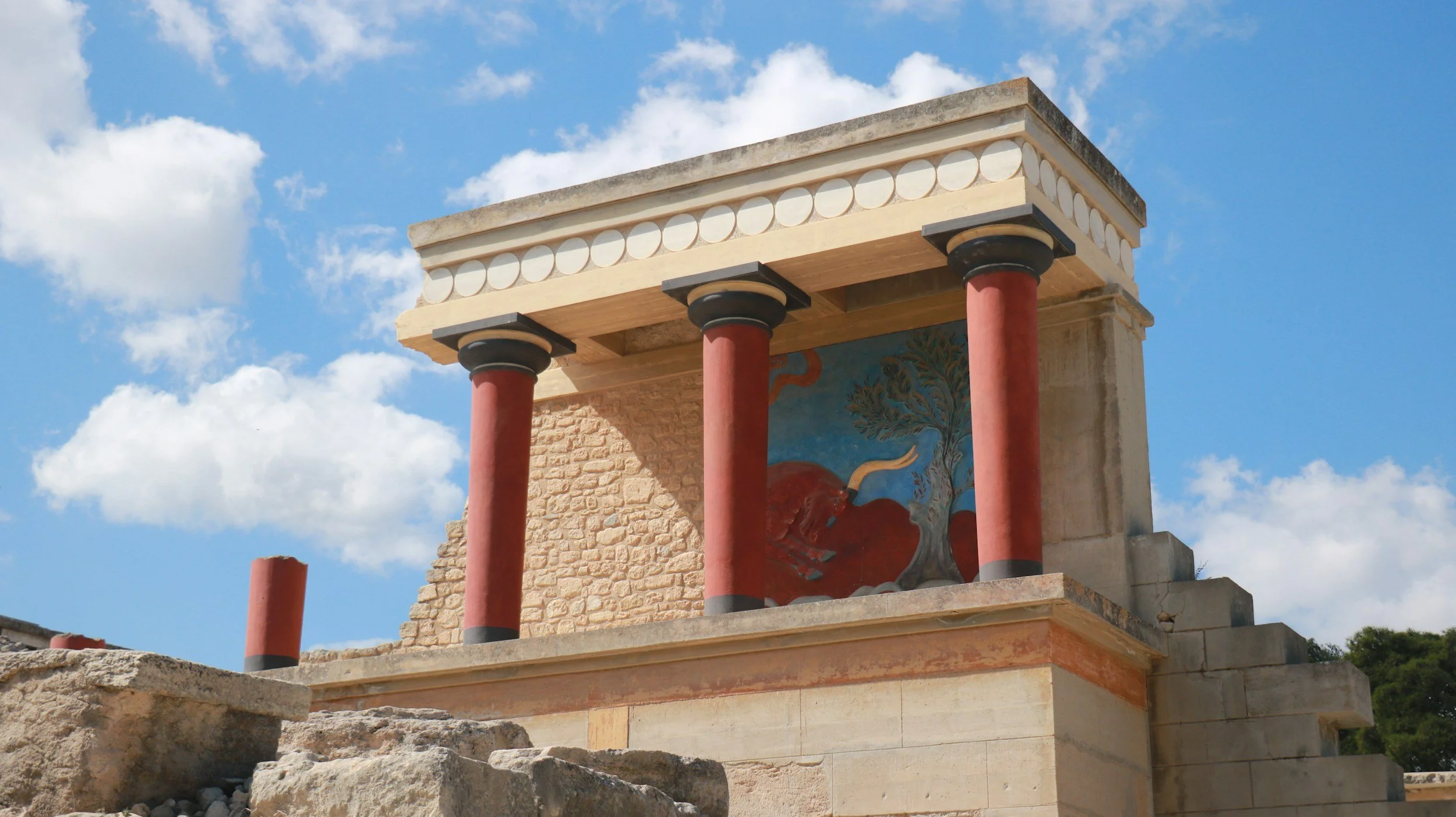 Ancient Greek-style ruin with red columns, decorative frieze, and painted mural of a bull and tree under a blue sky with clouds.