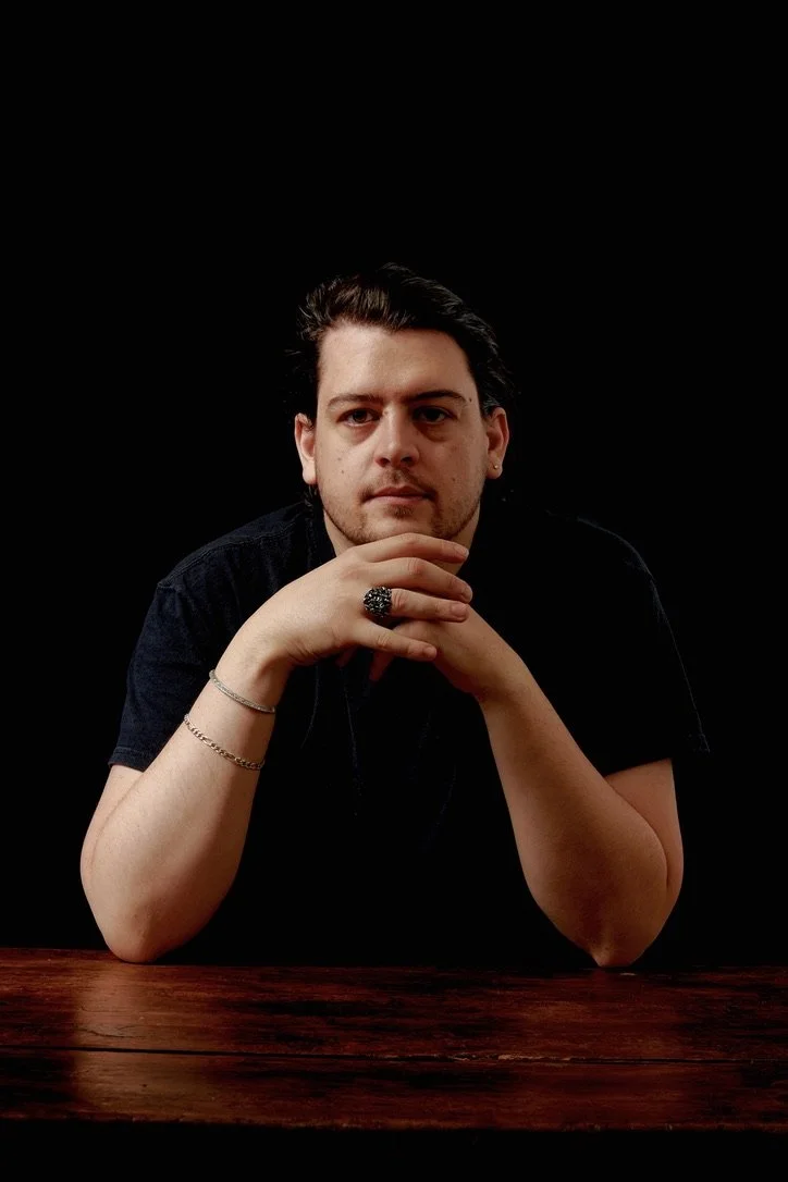 A young man with dark hair and facial hair, sitting at a wooden table with his hands clasped under his chin, wearing a black shirt and rings, against a dark background.