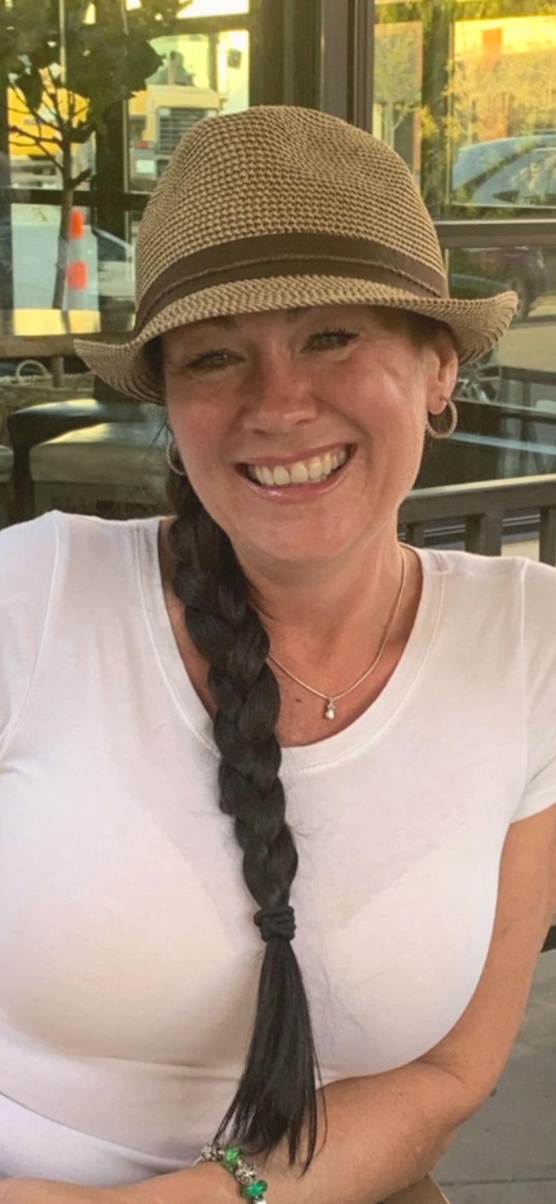 A woman wearing a tan fedora hat, white t-shirt, and a necklace, smiling while sitting indoors with a window behind her.