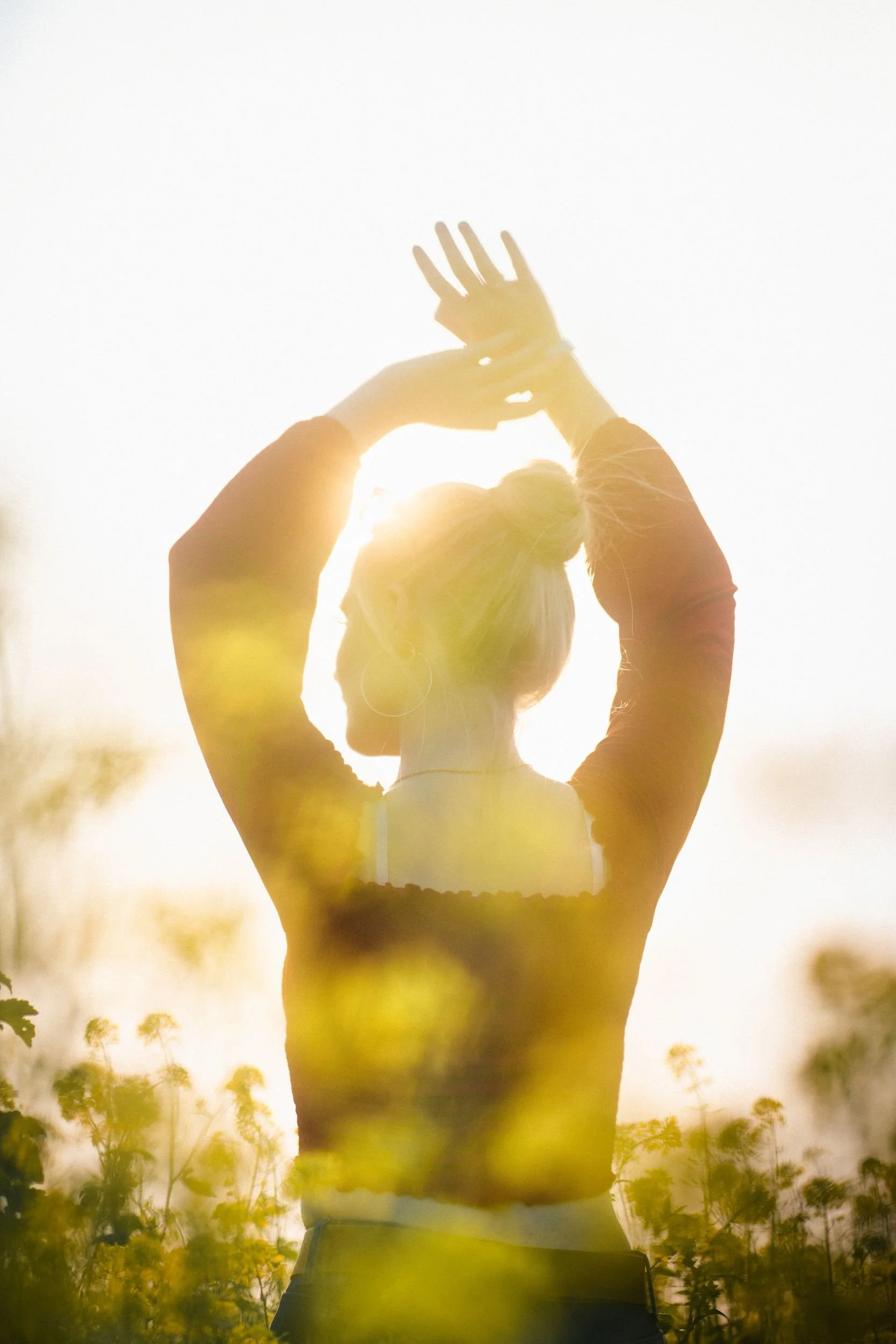 Person standing outdoors with arms raised, back and sunlit in a field of flowers with sunlight creating a warm glow.