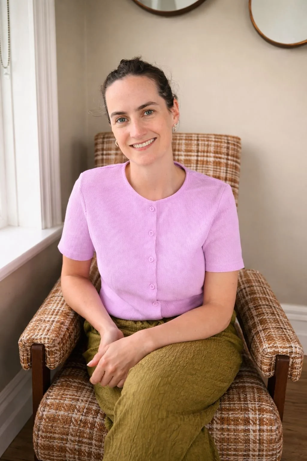 A woman with dark hair tied back, wearing a pink short-sleeved cardigan and olive-green pants, sitting in a vintage armchair with a checkered pattern, smiling at the camera, in a cozy room with beige walls and circular wall decor.