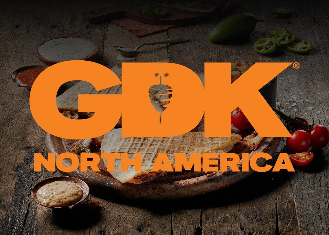 A wooden table holding a plate of grilled cheese sandwiches, cherry tomatoes, jalapeños, and dipping sauces, with a large orange GDK North America logo overlay.