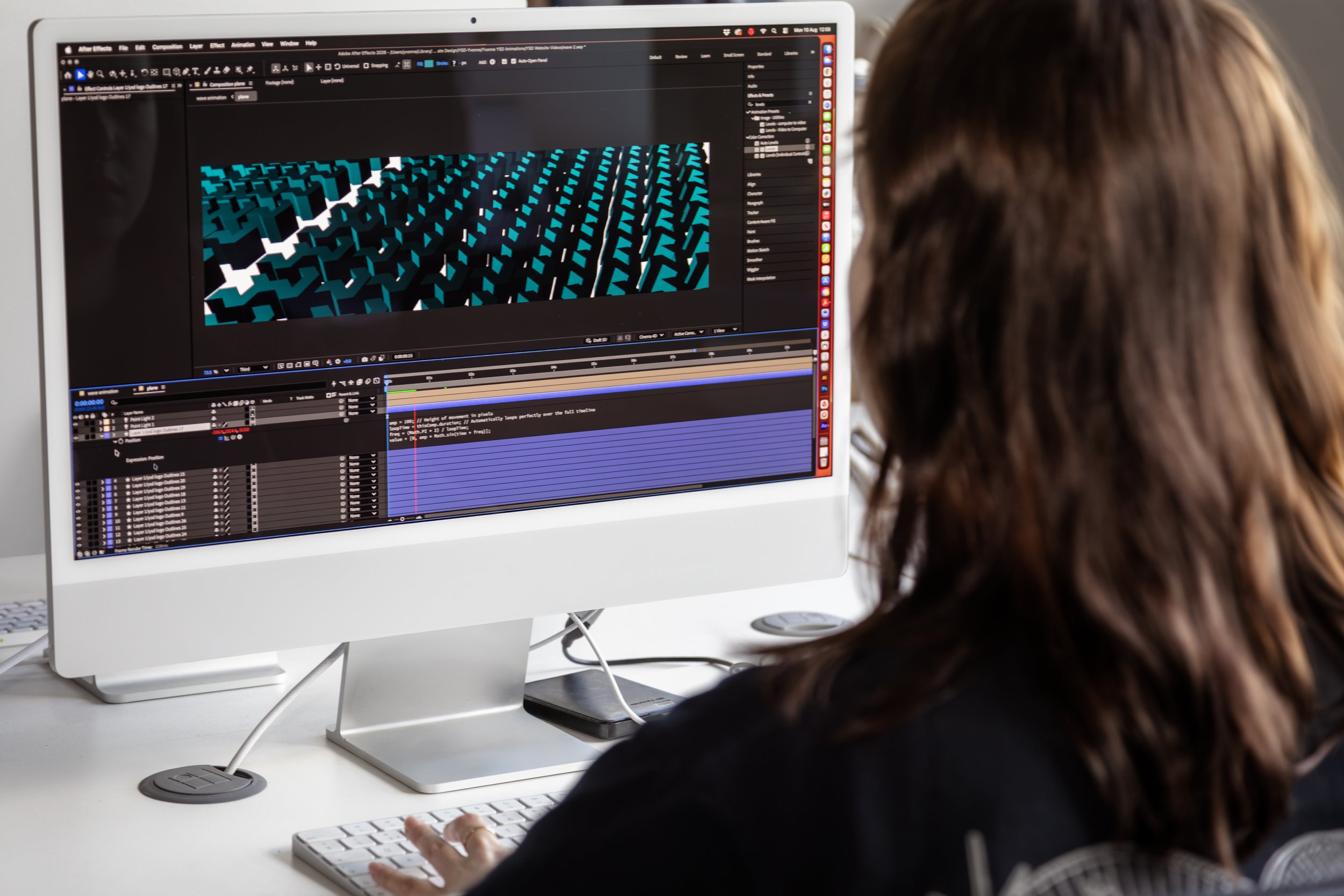 A person with brown hair working on video editing software on an iMac computer in a bright office environment.