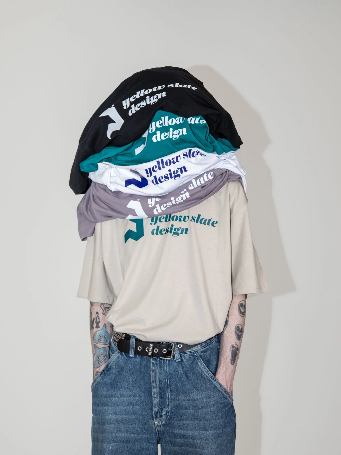 A person with tattoos on their arms, wearing a beige t-shirt, blue jeans, and a black belt, standing against a plain wall. Four folded t-shirts with the text 'yellow slate design' and a logo are draped over their shoulders.