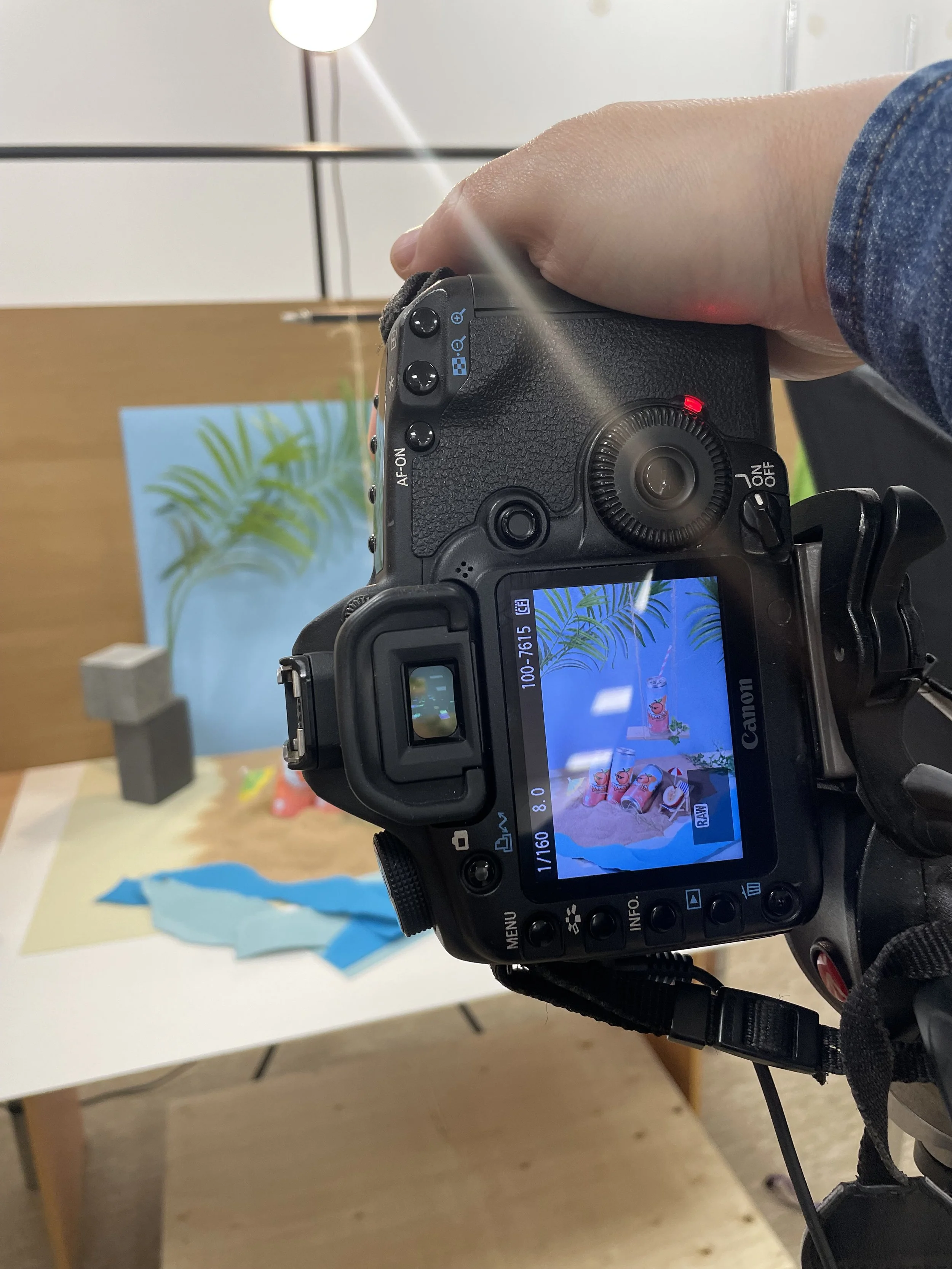 A digital camera capturing an image of colorful paper and objects on a table with a background of a blue poster featuring green leaves.