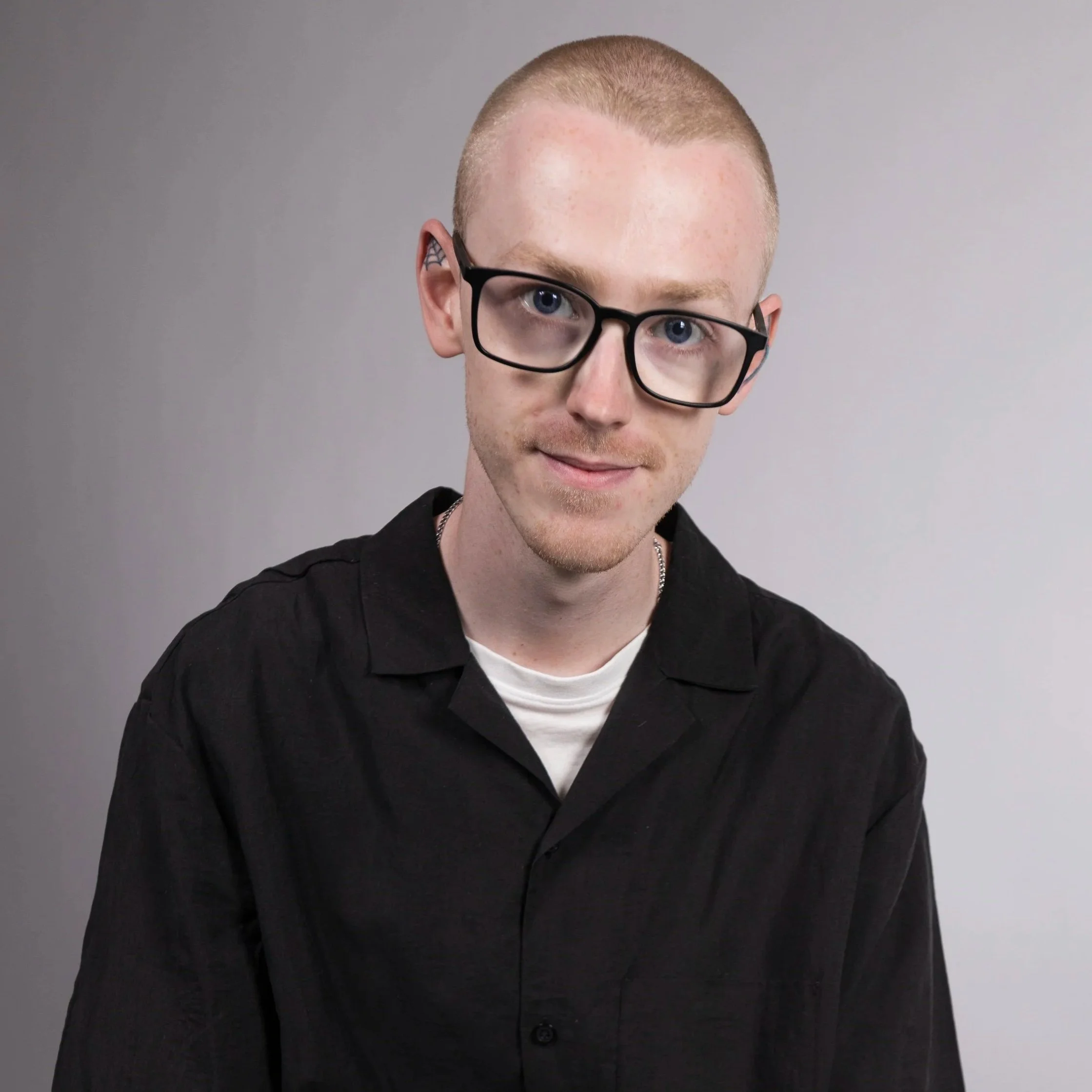 Close-up of a young man with short hair, wearing glasses, a black shirt, and a white undershirt, against a plain grey background.