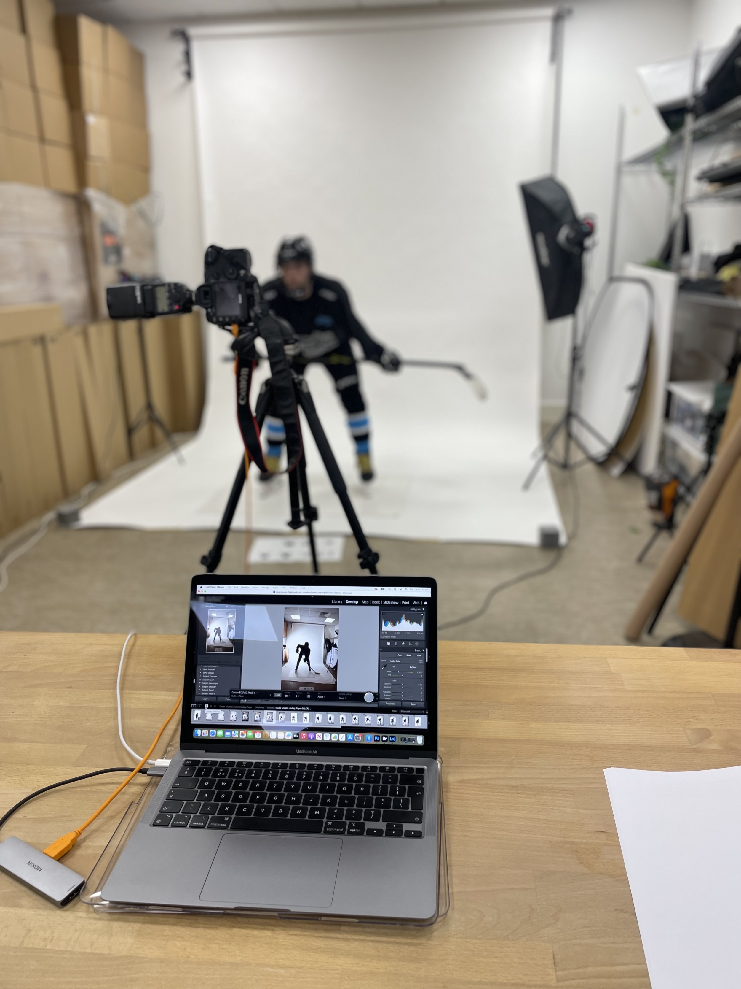 Photo of a photography studio with a MacBook Air laptop on a wooden table displaying an editing software interface, a connected external drive, and a sheet of paper. In the background, a professional camera on a tripod faces a white seamless backdrop where a hockey player in uniform is posing with a hockey stick. Studio lighting and shelves with storage boxes are also visible.