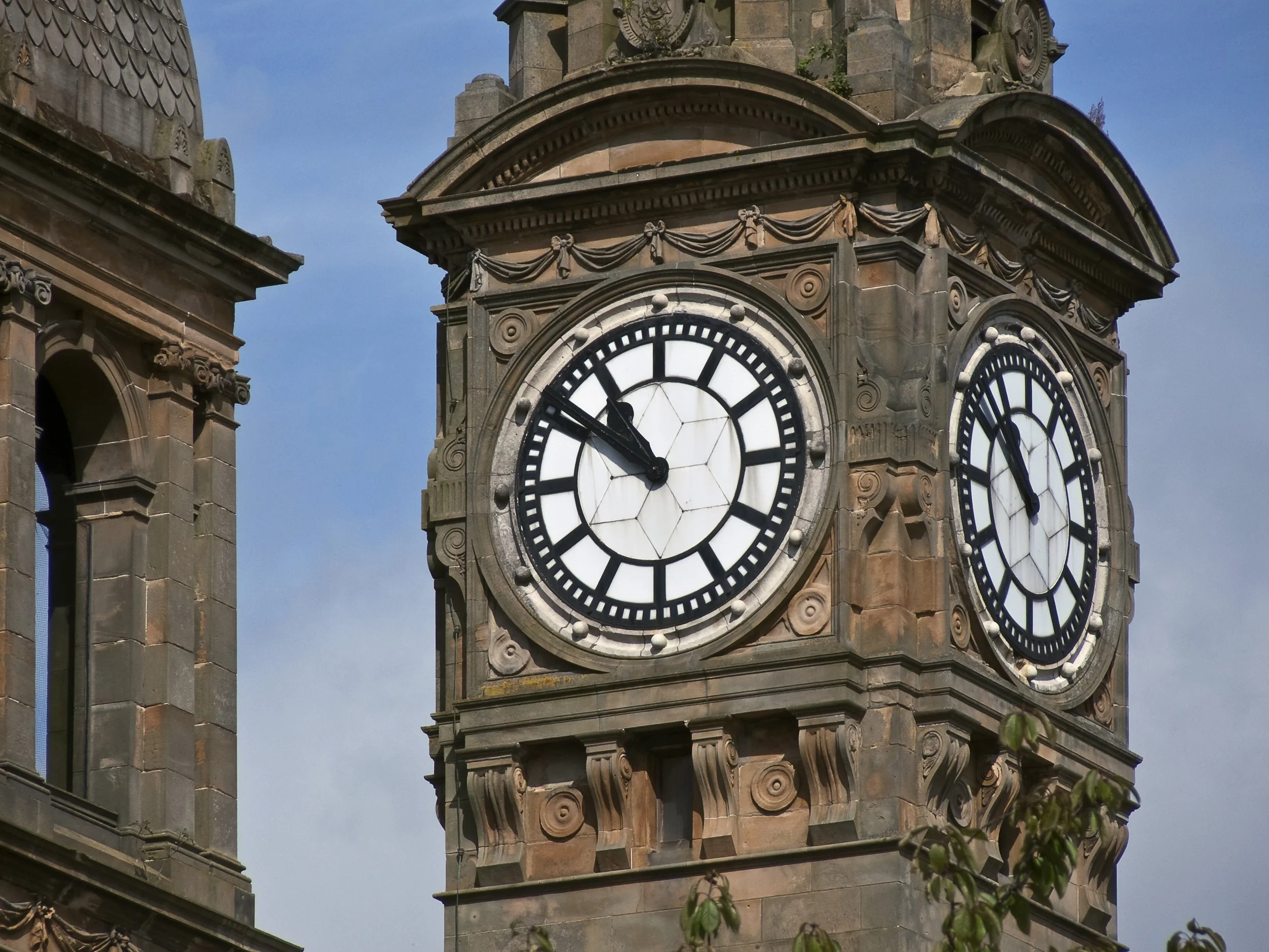 Close-up of an ornate, historic clock tower made of stone, featuring decorative carvings and a large clock face with Roman numerals, showing the time as approximately 4:55.