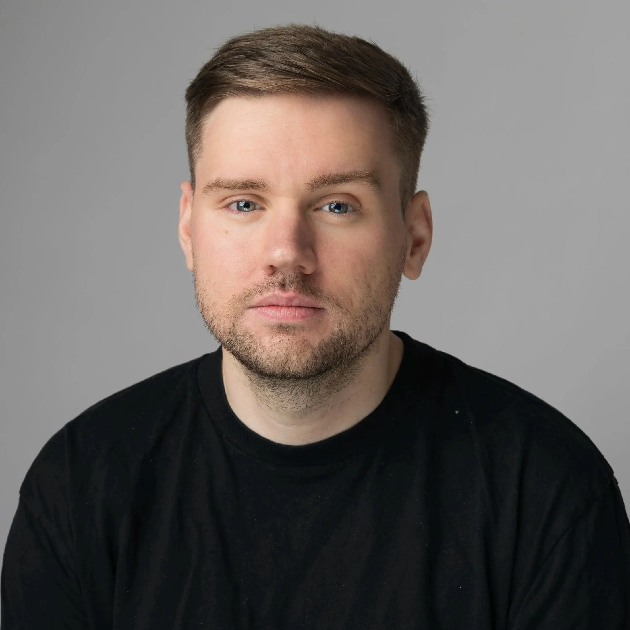 Close-up of a young man with brown hair, blue eyes, and light skin, wearing a black shirt against a plain gray background.
