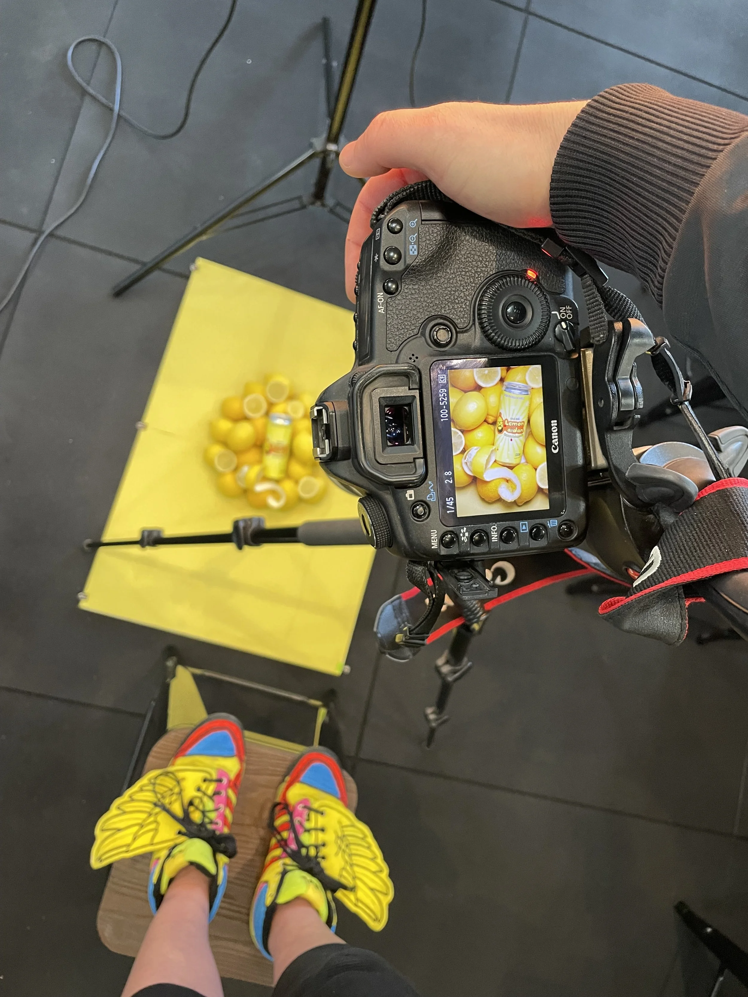 A person wearing yellow sneakers with pink, black, and blue accents standing on a skateboard, taking a photo of a yellow lemons display with a camera. The camera screen shows a close-up of lemons and lemon juice bottles, with a yellow background.