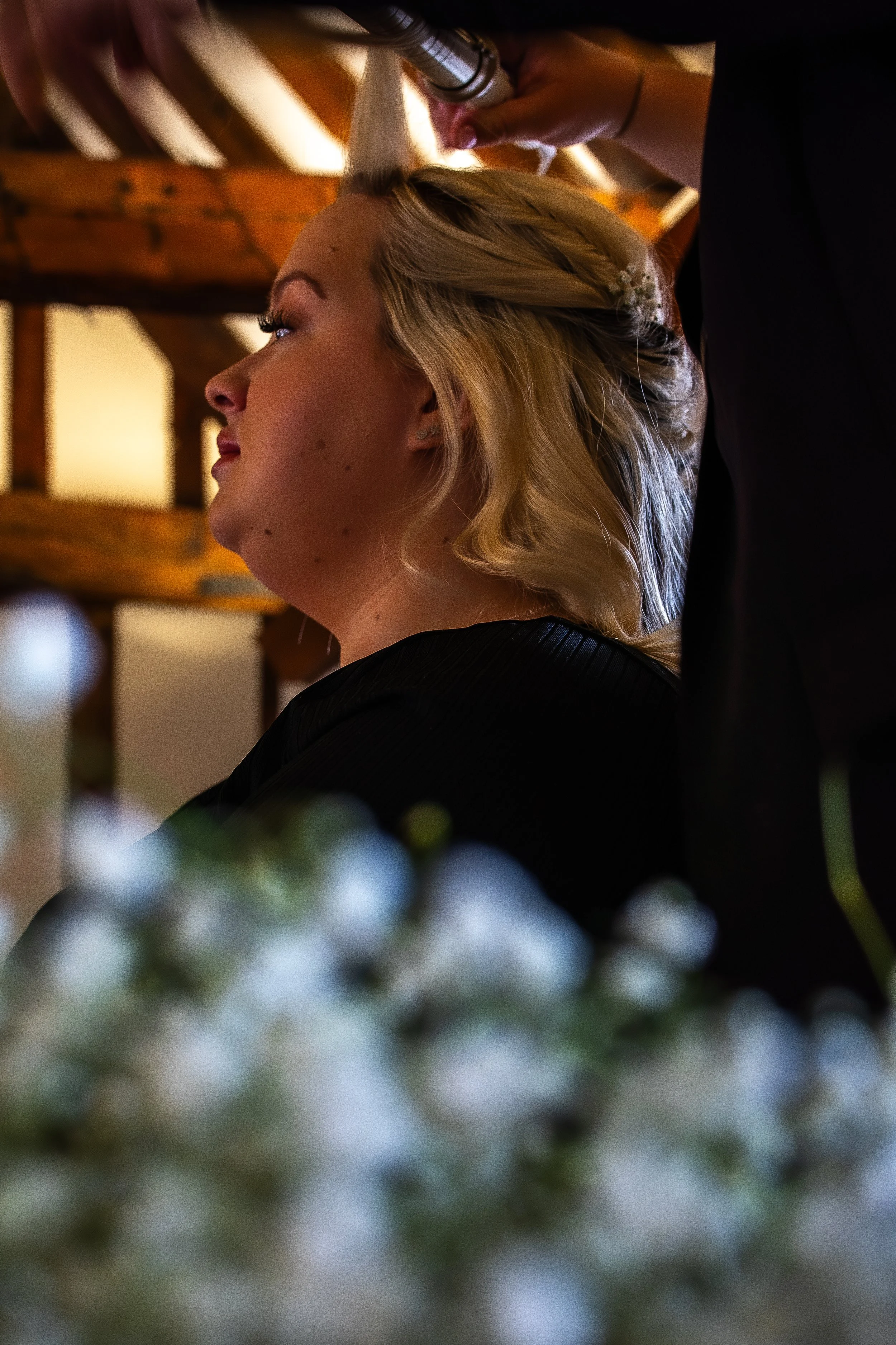 A woman with blonde hair getting her hair styled or paid in a salon setting, with a stylist working on her hair, and blurred flowers in the foreground.