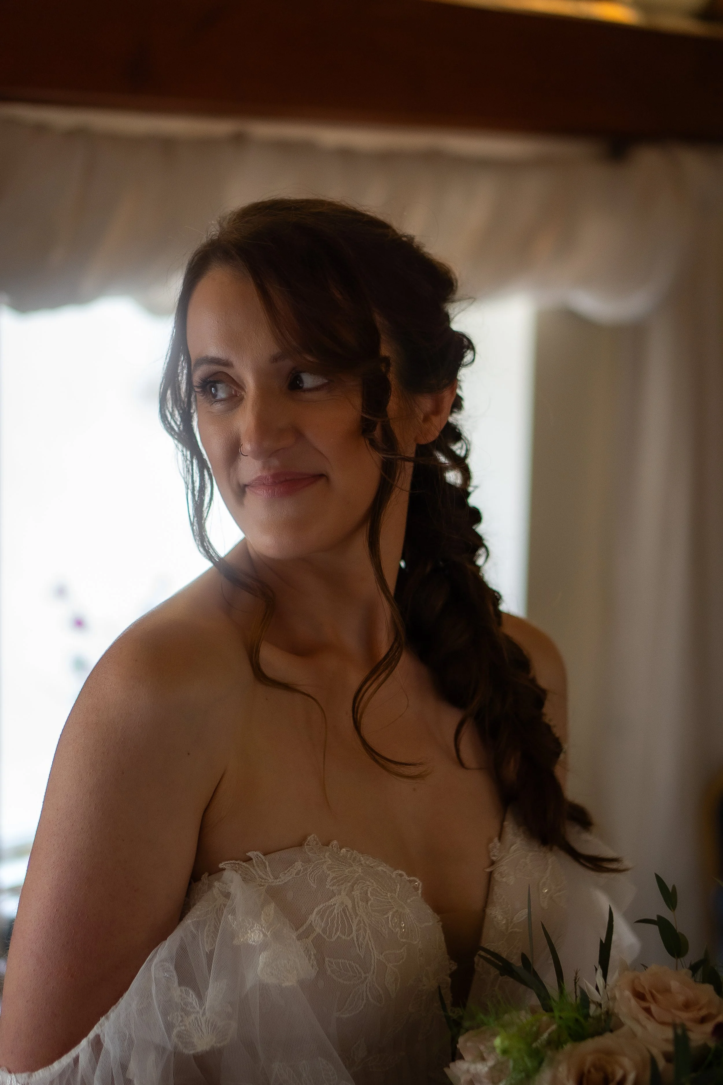 A woman with long, curly brown hair styled in an elegant braid, wearing a lace white dress, standing indoors near a window with a floral arrangement in the foreground.