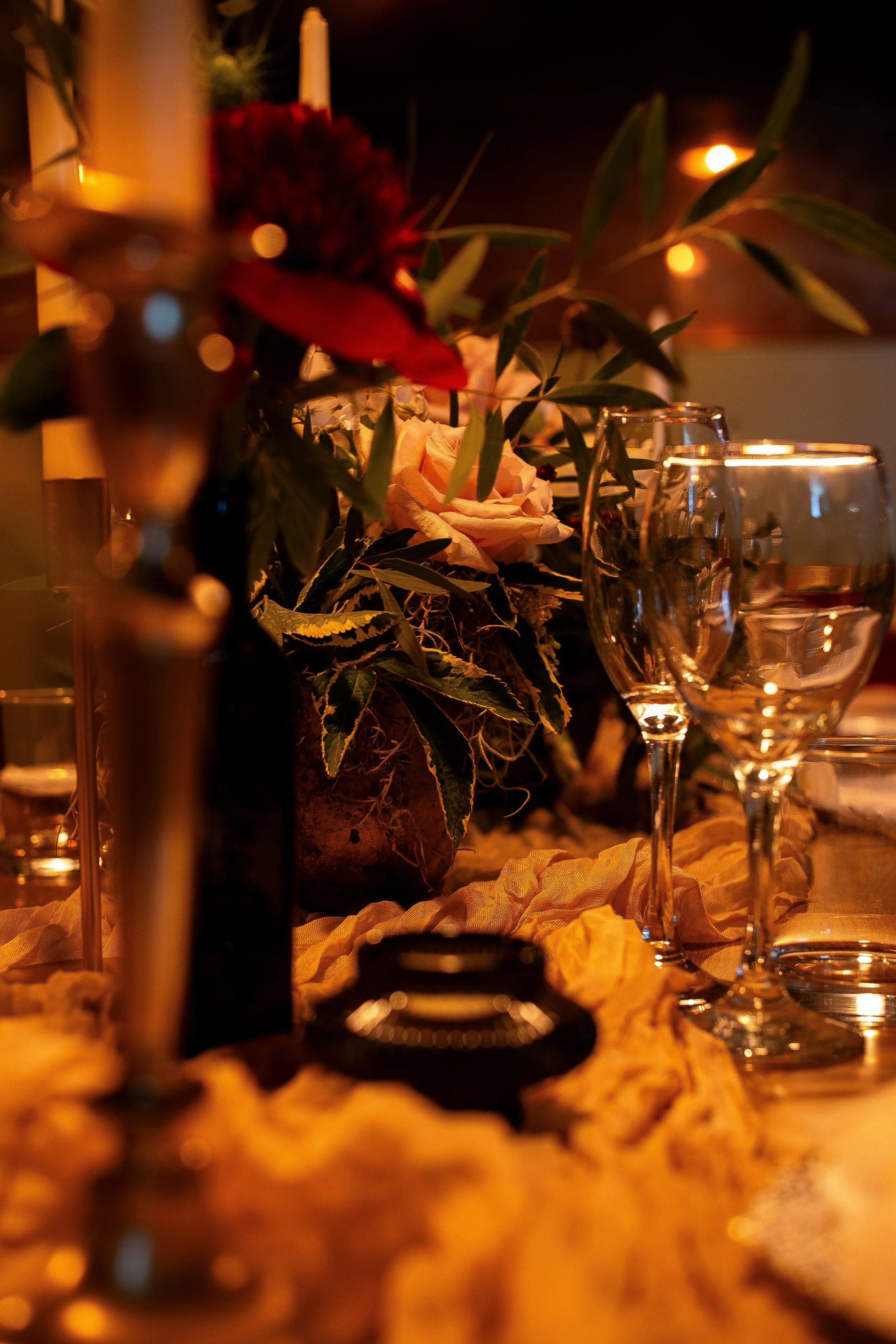 Close-up of a table centerpiece with flowers and wine glasses, illuminated by warm candlelight at a dinner event.