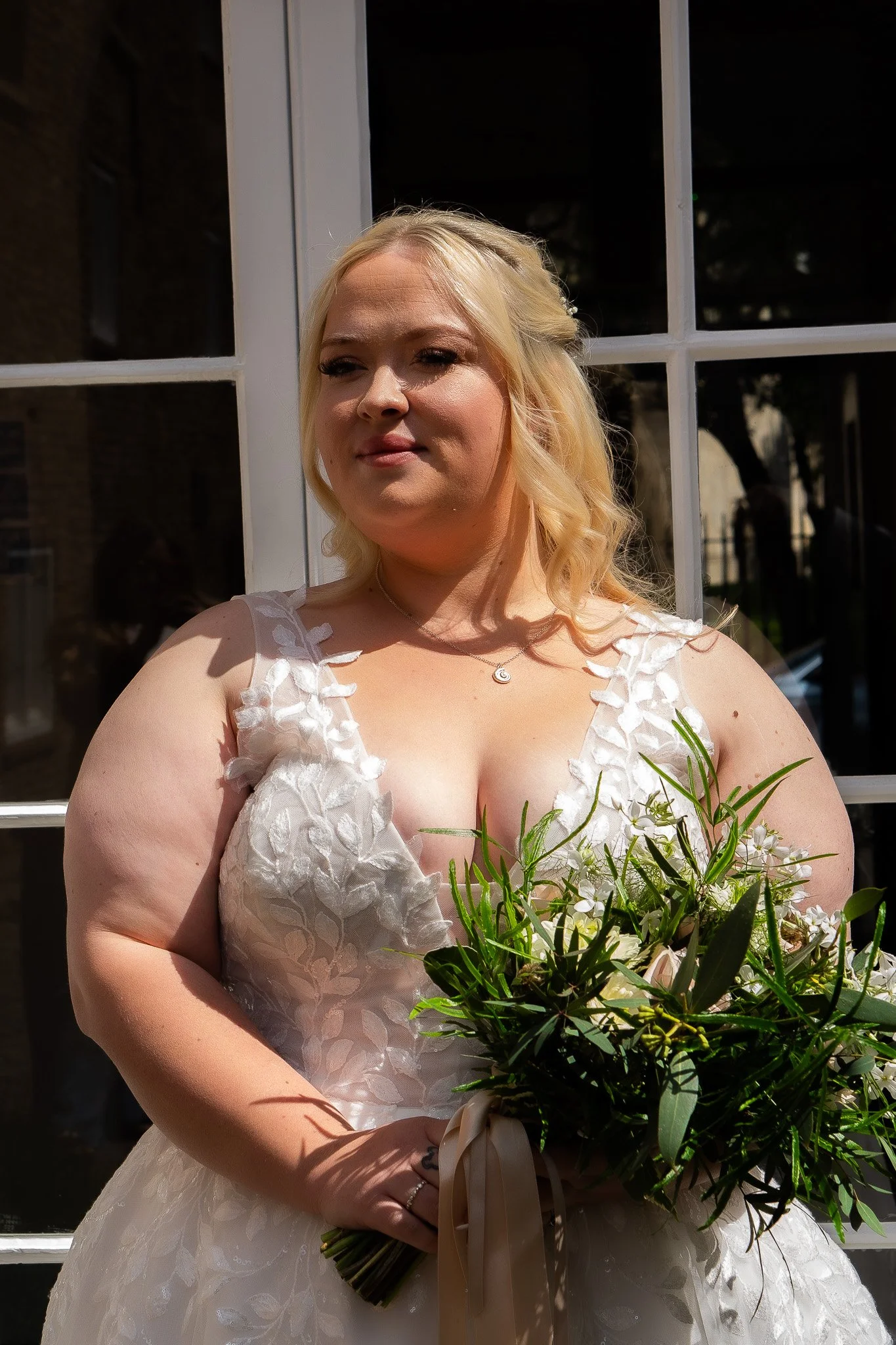 A woman in a white wedding dress holding a bouquet of green leaves and white flowers, standing outside in front of a window.