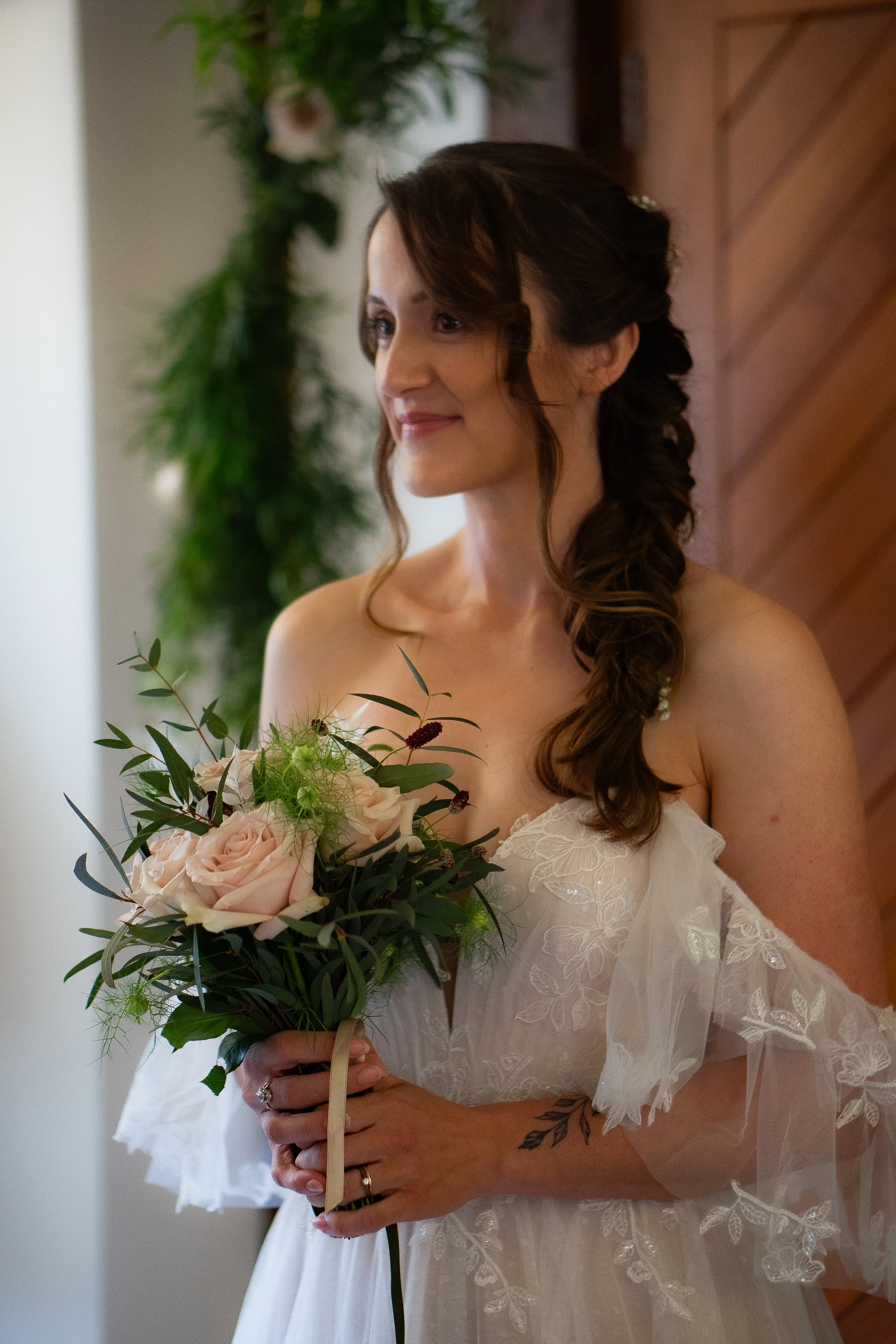 A woman dressed in a wedding dress holding a bouquet of pink and green flowers, standing indoors near a wooden wall. Her hair is styled in loose curls, and she has a slight smile.