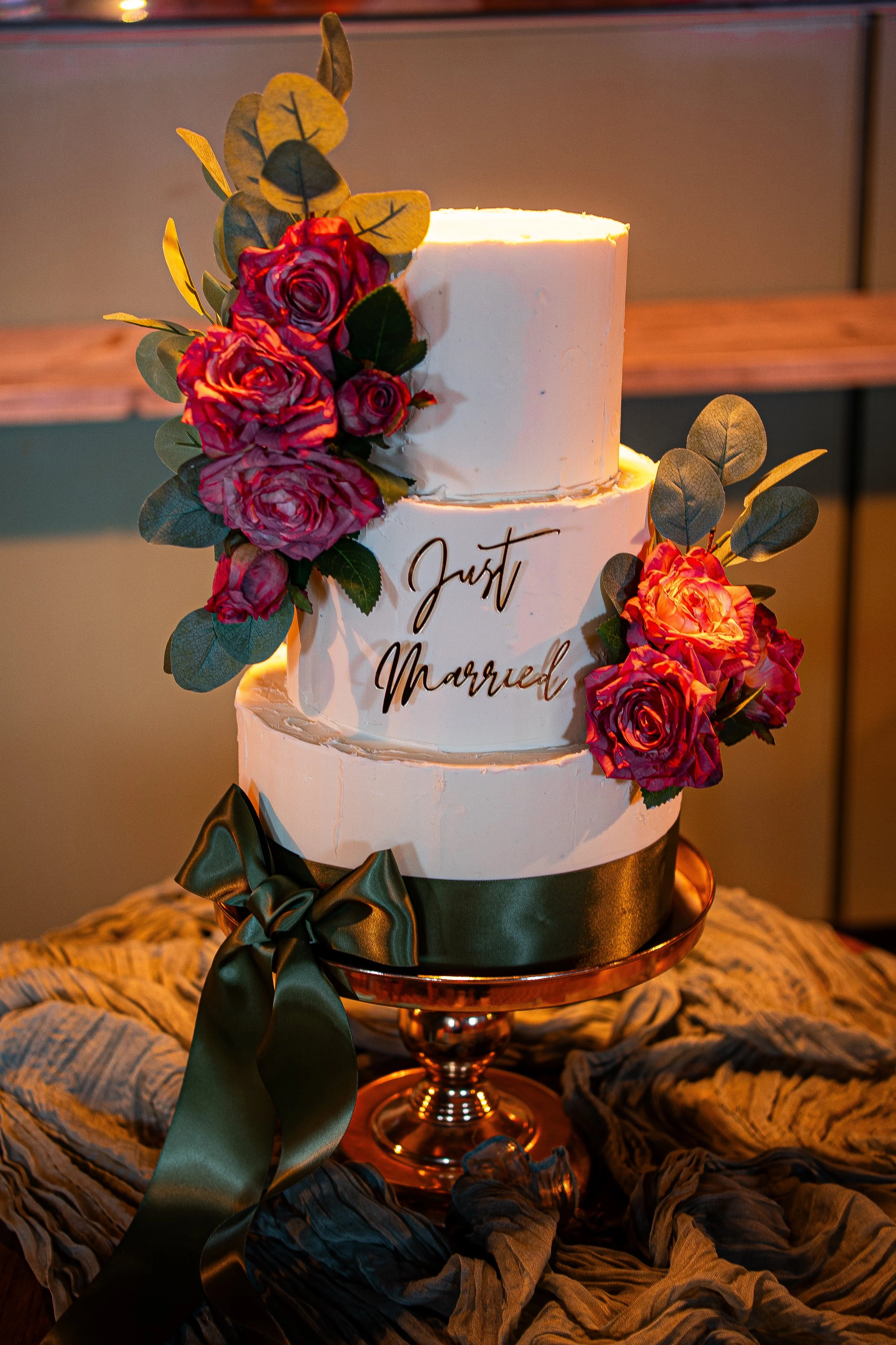 A three-tiered wedding cake decorated with pinkish-red roses, green leaves, and a dark green satin ribbon tied into a bow at the bottom, displayed on a golden cake stand. The cake has the words "Just Married" written on the middle tier.