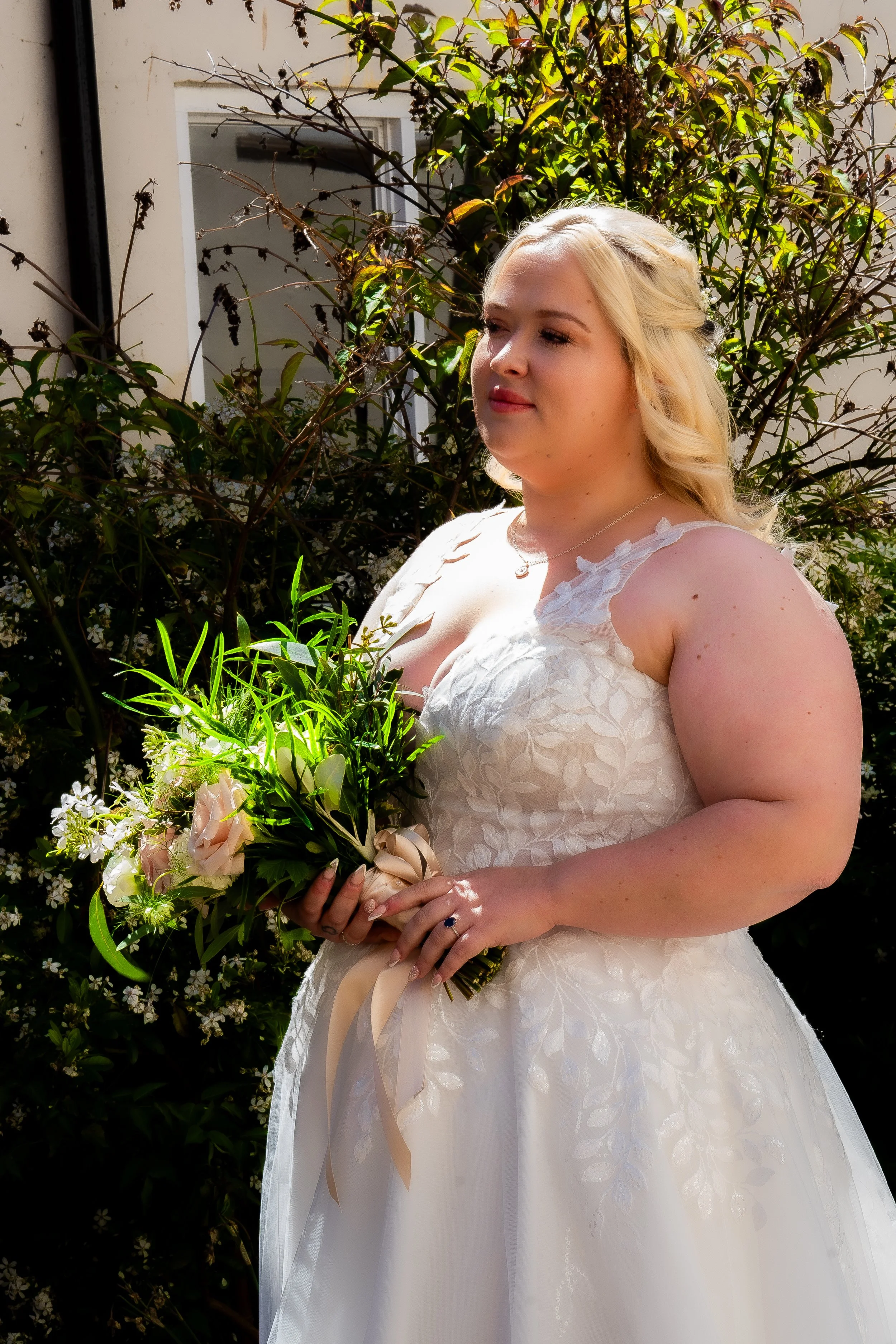 A woman in a wedding dress holding a bouquet of pink roses and greenery, standing outdoors in front of a bush with green and brown leaves.
