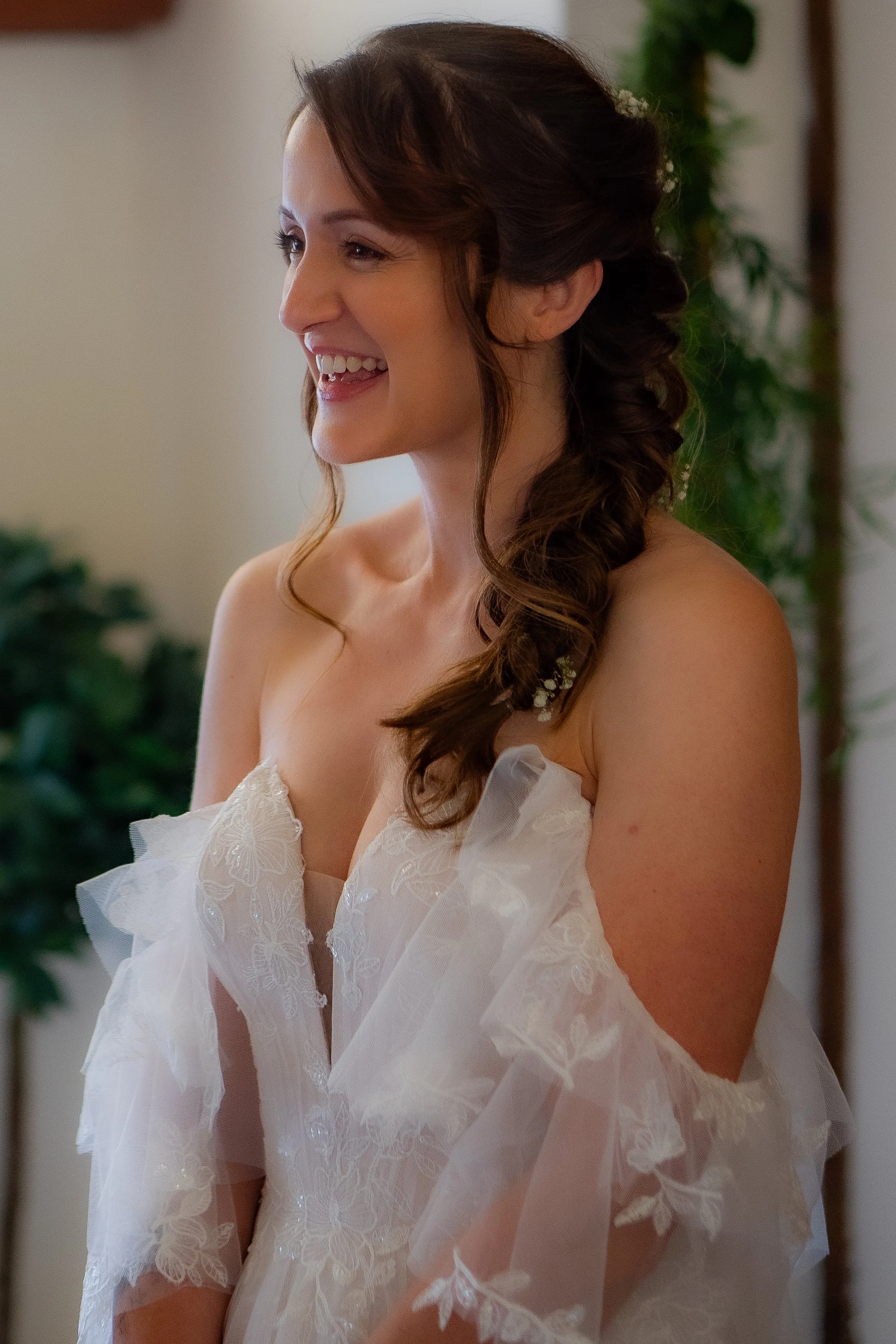 A woman with long brown hair styled in soft waves, wearing a white lace dress with floral embroidery, is smiling and looking to her left. She appears to be indoors, with green plants and a neutral-colored wall in the background.