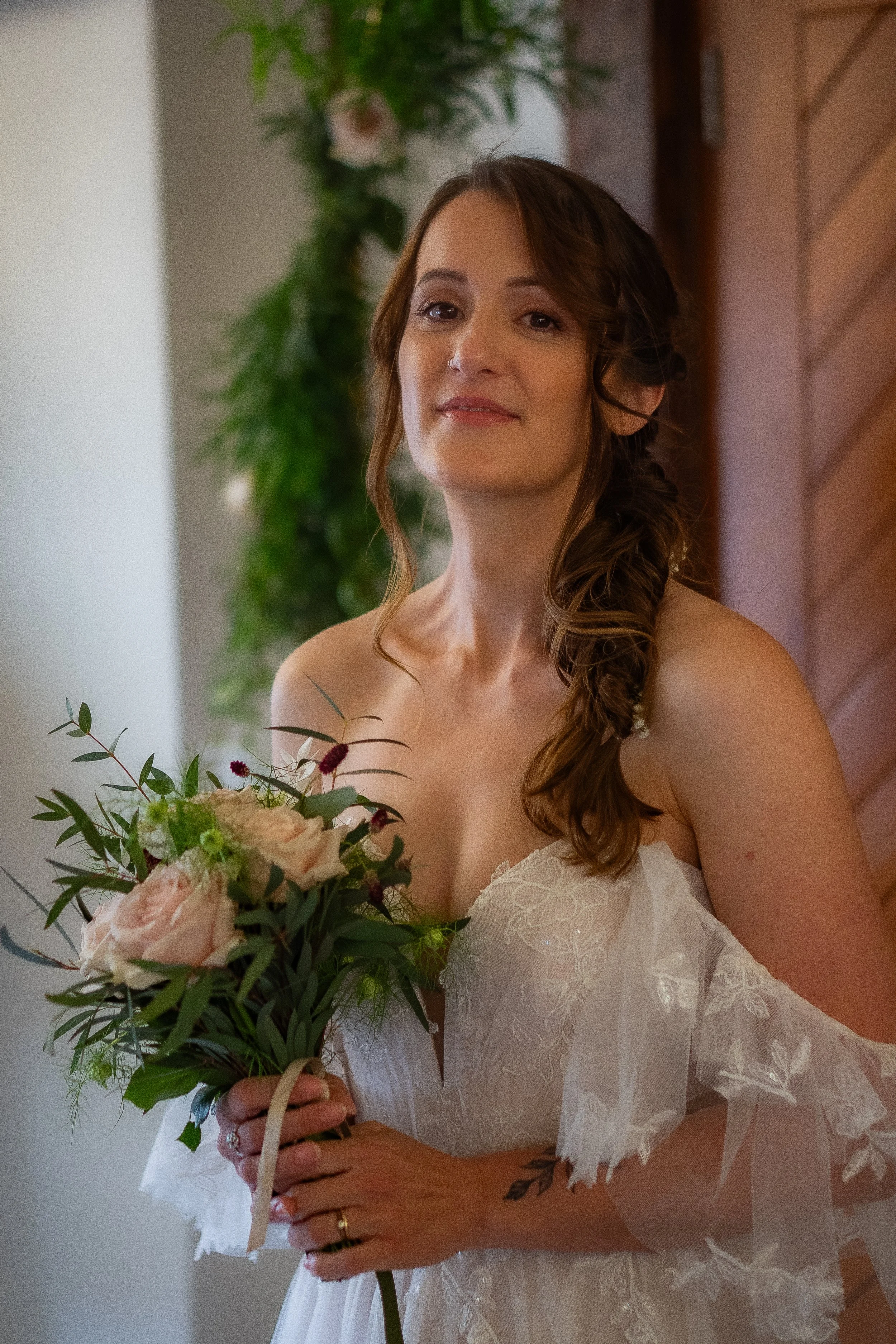 A woman in a white wedding dress holding a bouquet of pink roses, beige flowers, and eucalyptus leaves, with a wooden wall and green plants in the background.