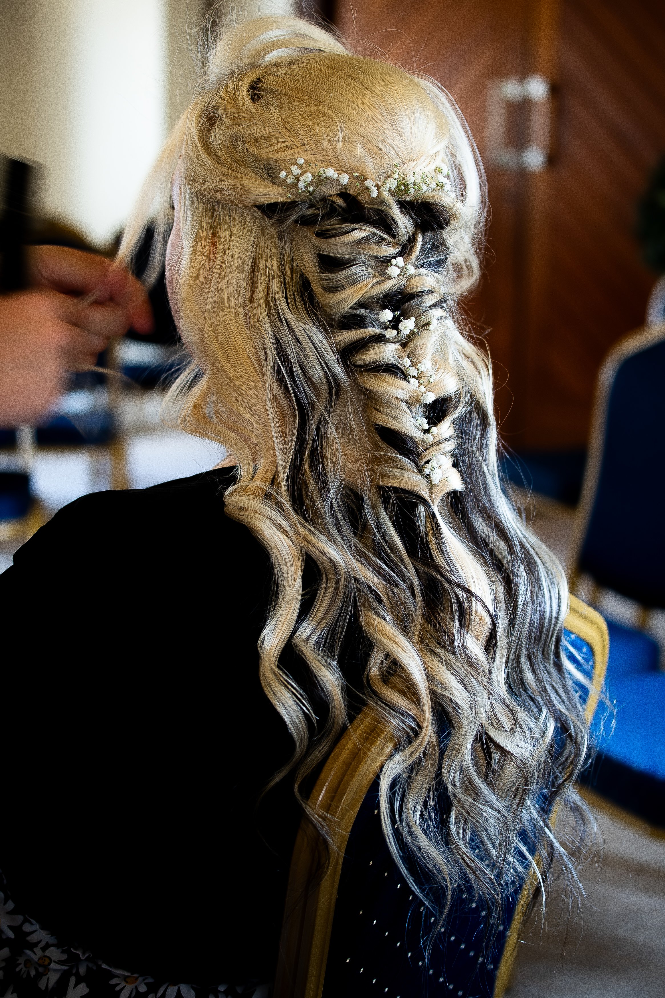 A woman with blonde hair styled in loose curls and small white flowers woven into her hair. A hand is visible, adjusting her hair, with a wooden chair and a brown wooden wall in the background.