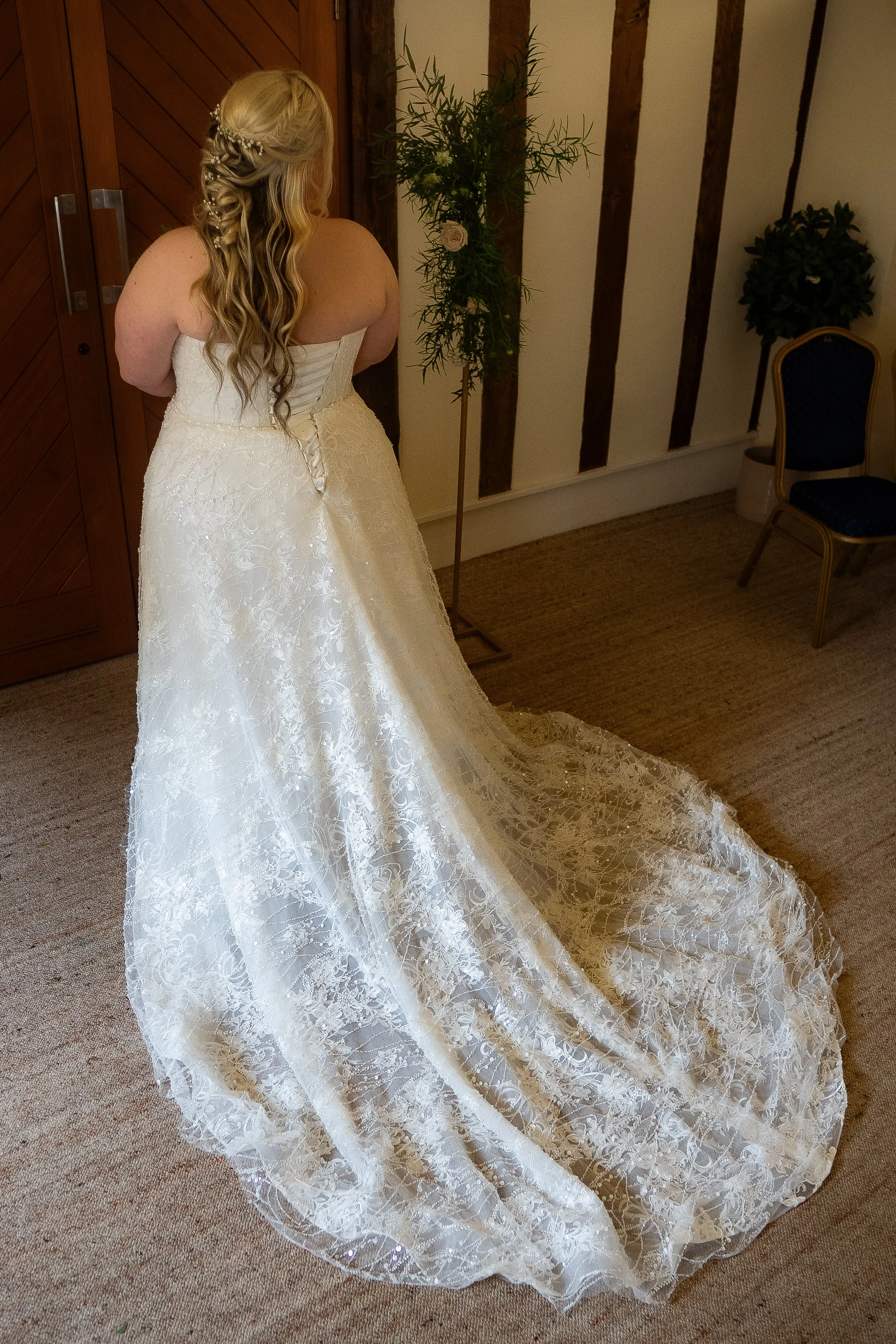 A woman in a lace wedding gown with a long train is standing with her back to the camera, facing a wooden wall with vertical wall beams and a floral arrangement.