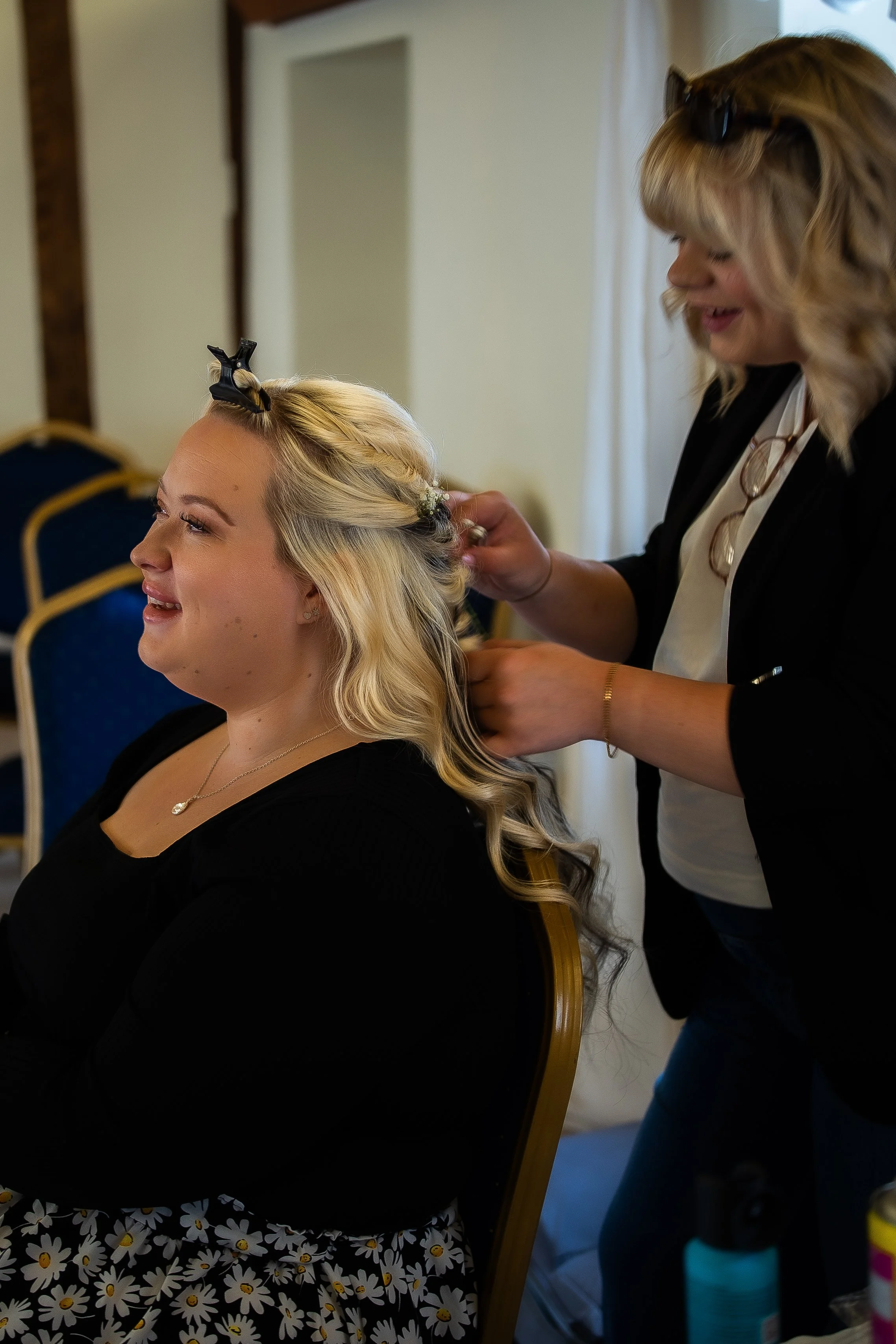A woman with blonde hair styled in loose curls is smiling while having her hair curled by another woman with blonde hair and sunglasses on her head. They are indoors, with chairs and miscellaneous items around.