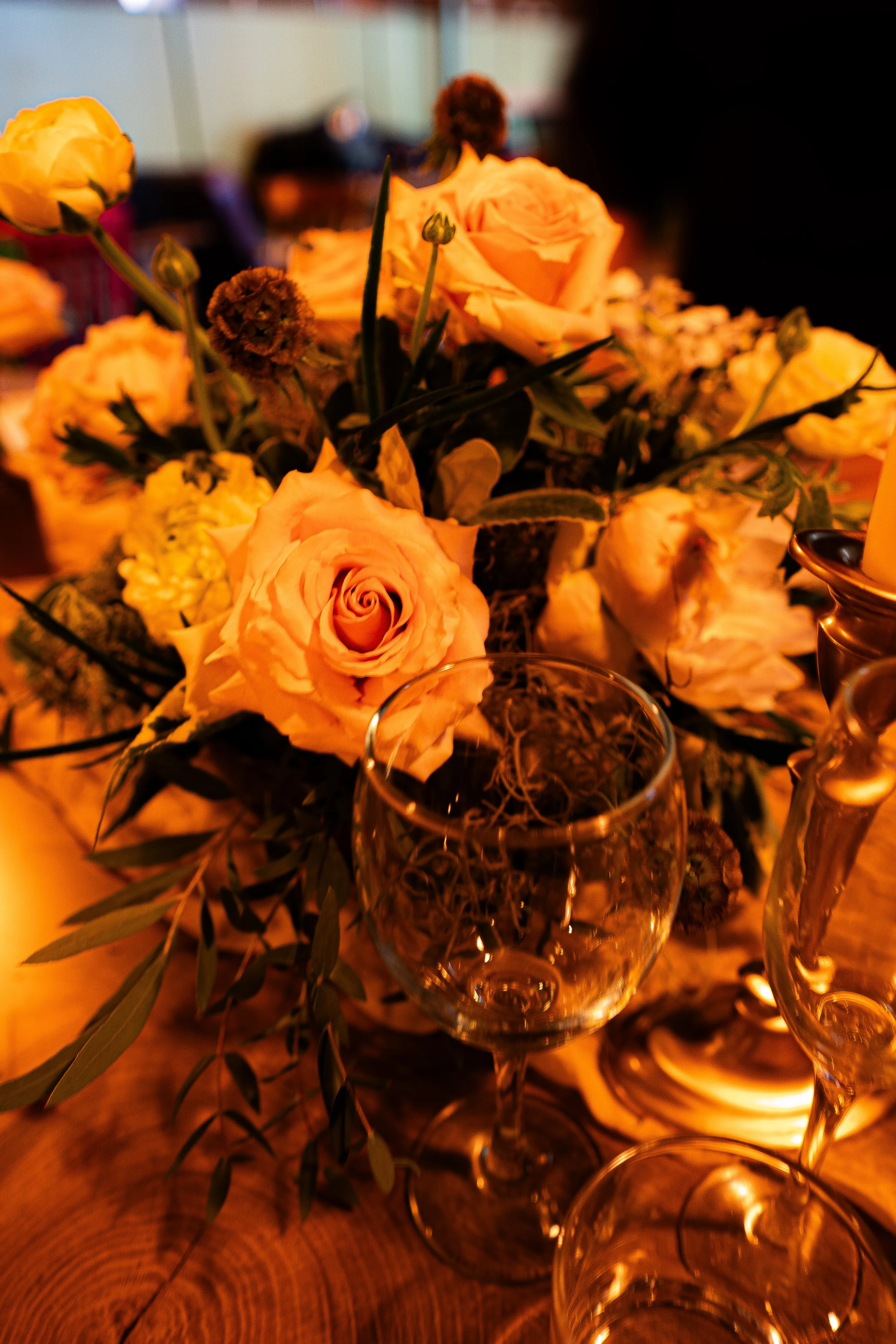 A close-up of a bouquet of yellow and peach roses with some greenery, placed inside a glass vase, with a warm, dimly lit background.