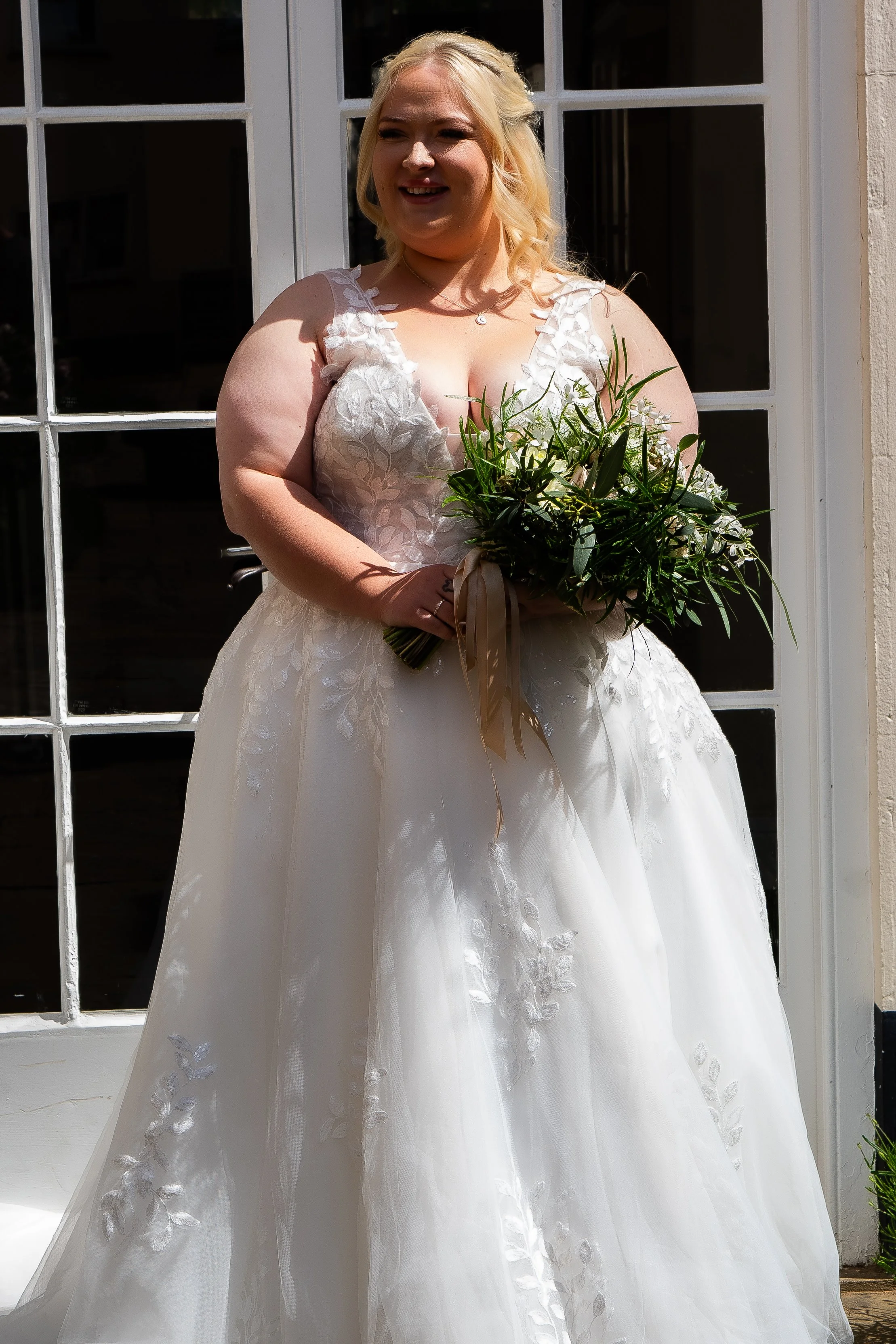 A woman in a white wedding dress holding a bouquet of greenery standing outside in front of a glass door with grid panes.