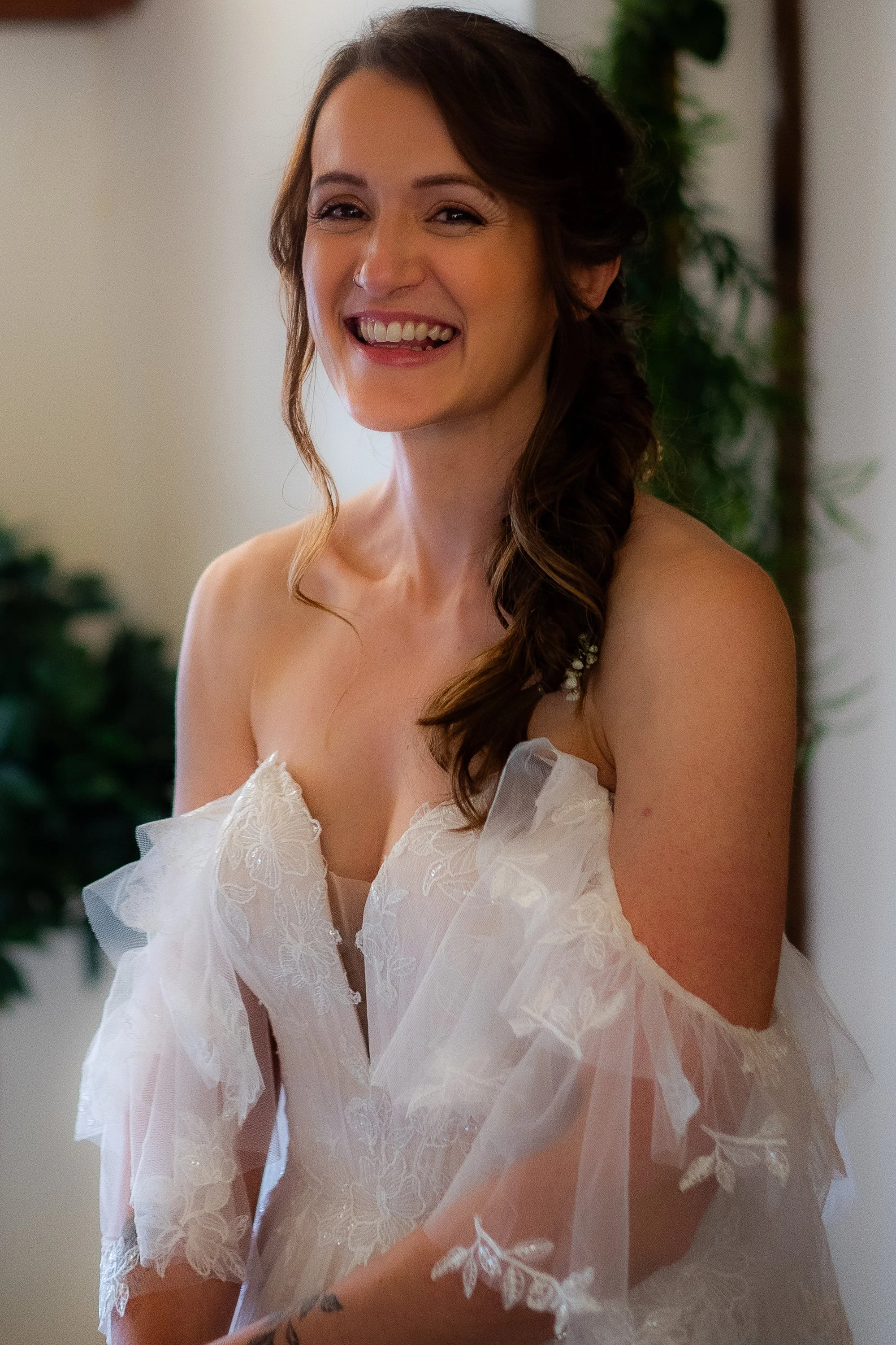 A woman with curly brown hair smiling, wearing a white lace dress with floral embroidery, standing indoors with a blurred background of green plants.