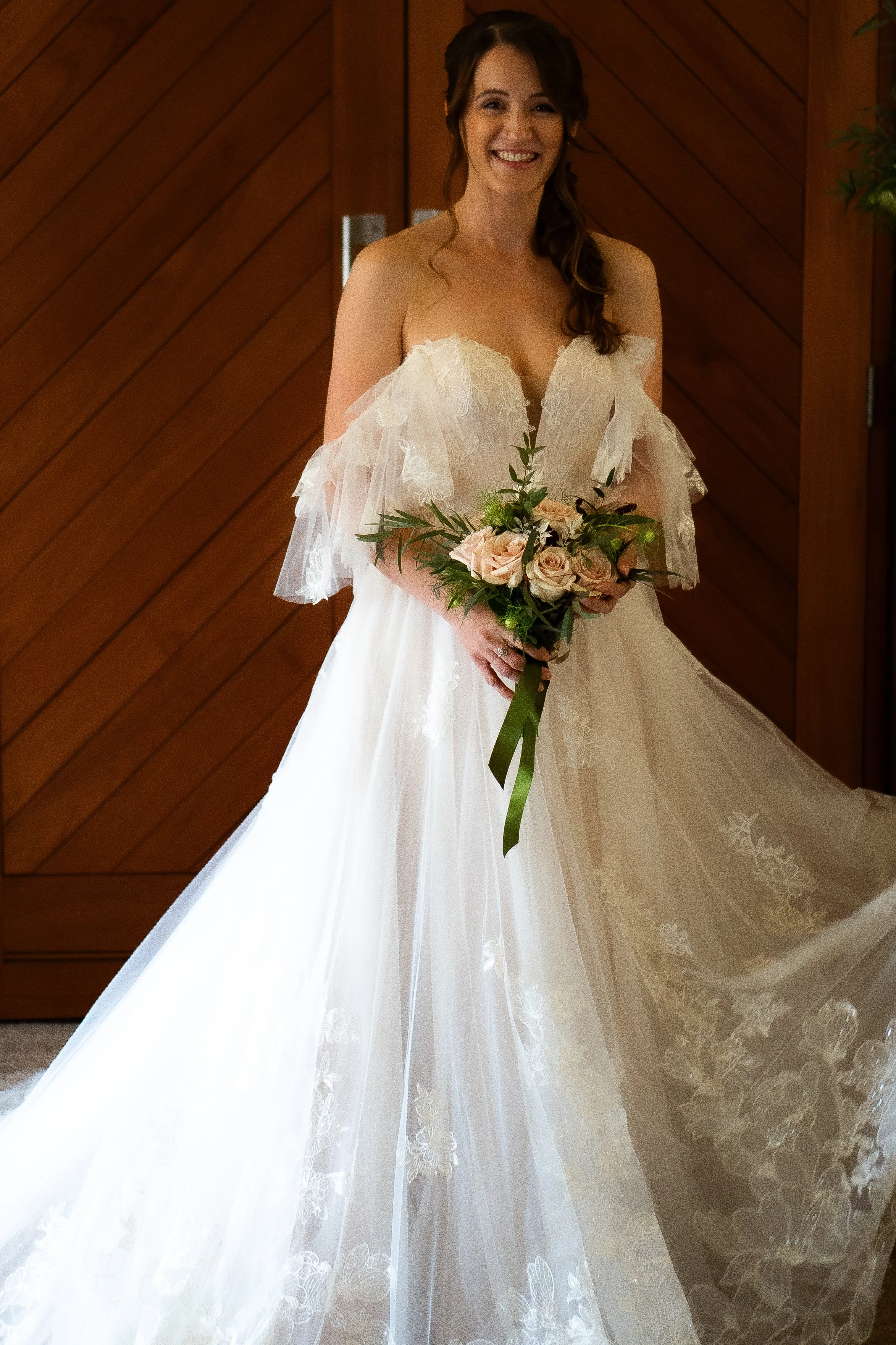 A woman in a wedding dress holding a bouquet of pink roses, standing indoors in front of a wooden wall, smiling.