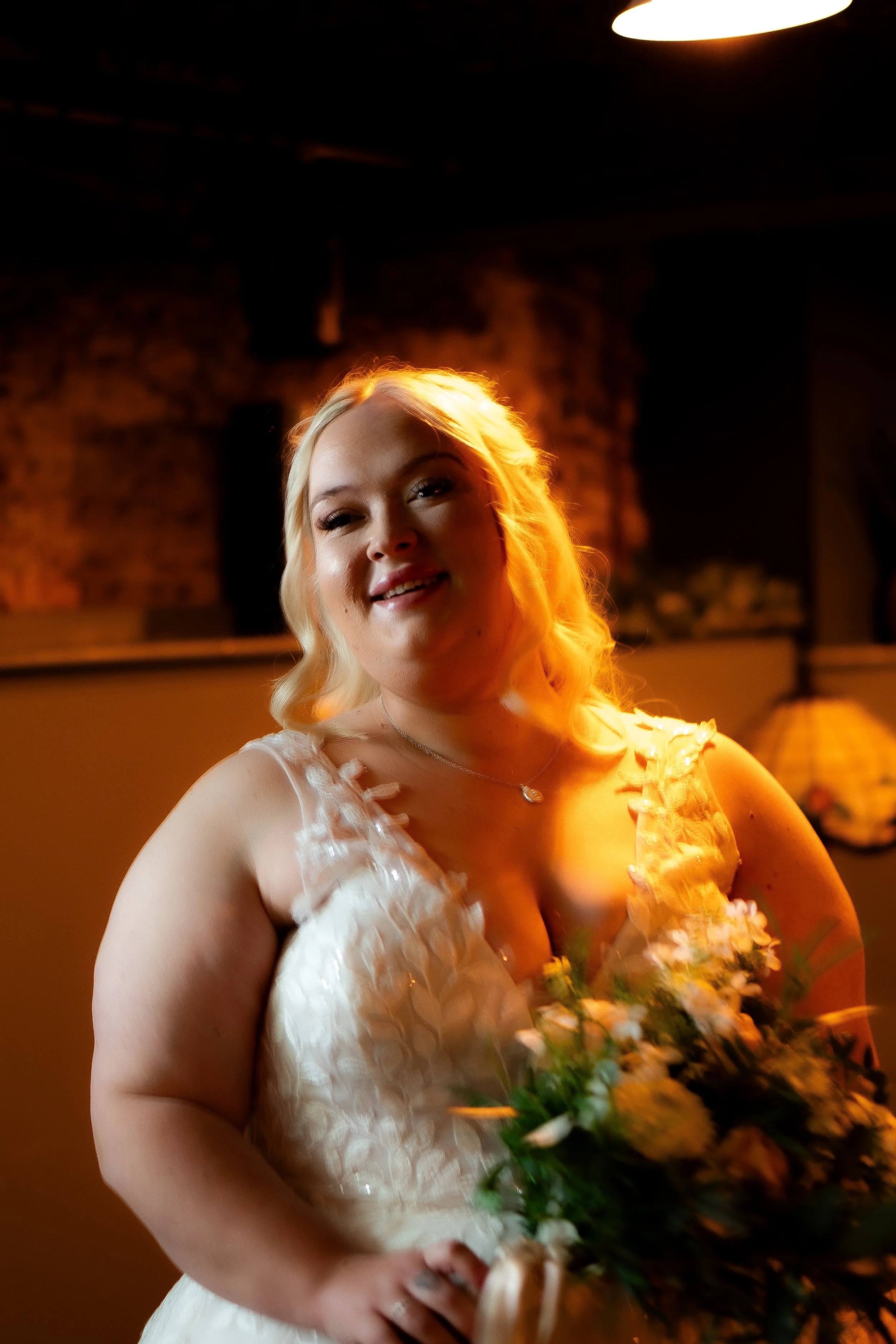 A young woman in a white dress holding a bouquet of flowers, smiling indoors with warm lighting and a brick wall background.